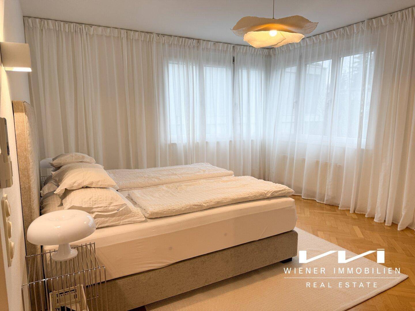 Gemütliches Schlafzimmer mit großem Bett, hellen Vorhängen und Parkettboden.