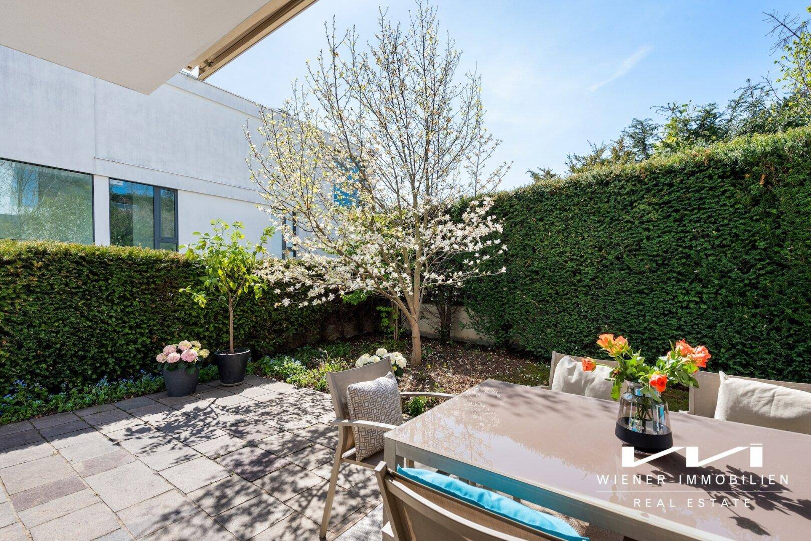Sonniger Terrassenbereich mit Esstisch, umgeben von Rasen, Hecken und blühenden Bäumen.