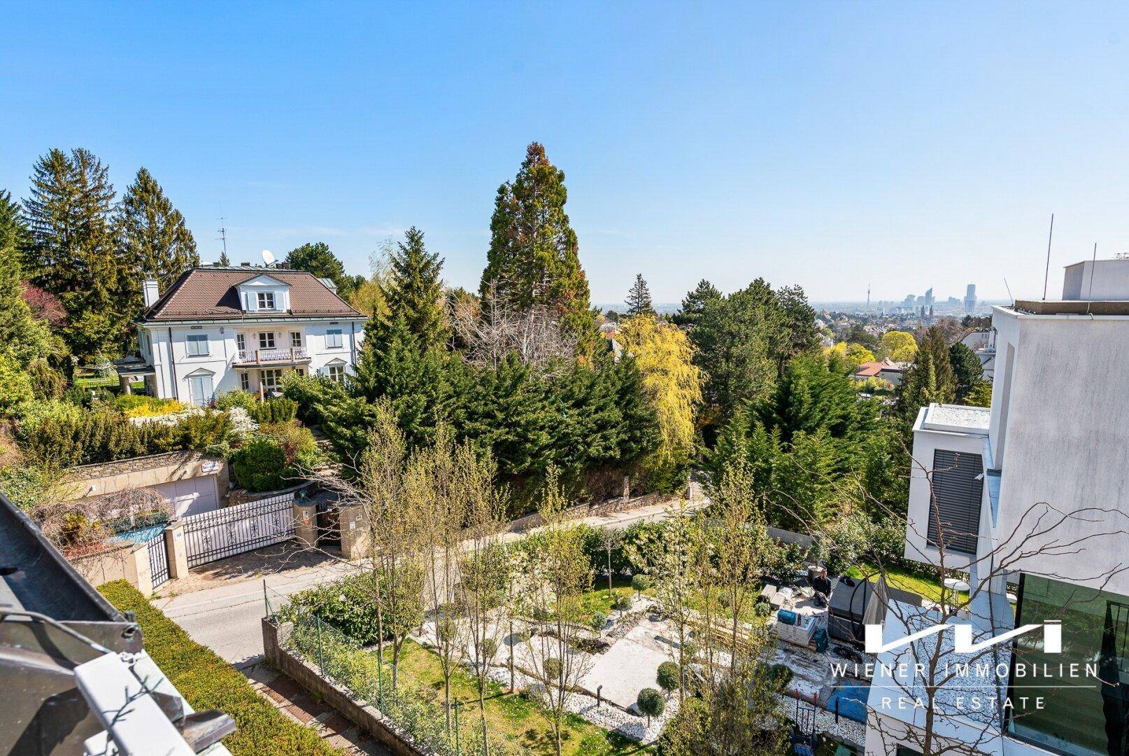 Weitläufiger Blick über die grüne Umgebung mit Villen und der Skyline der Stadt im Hintergrund.