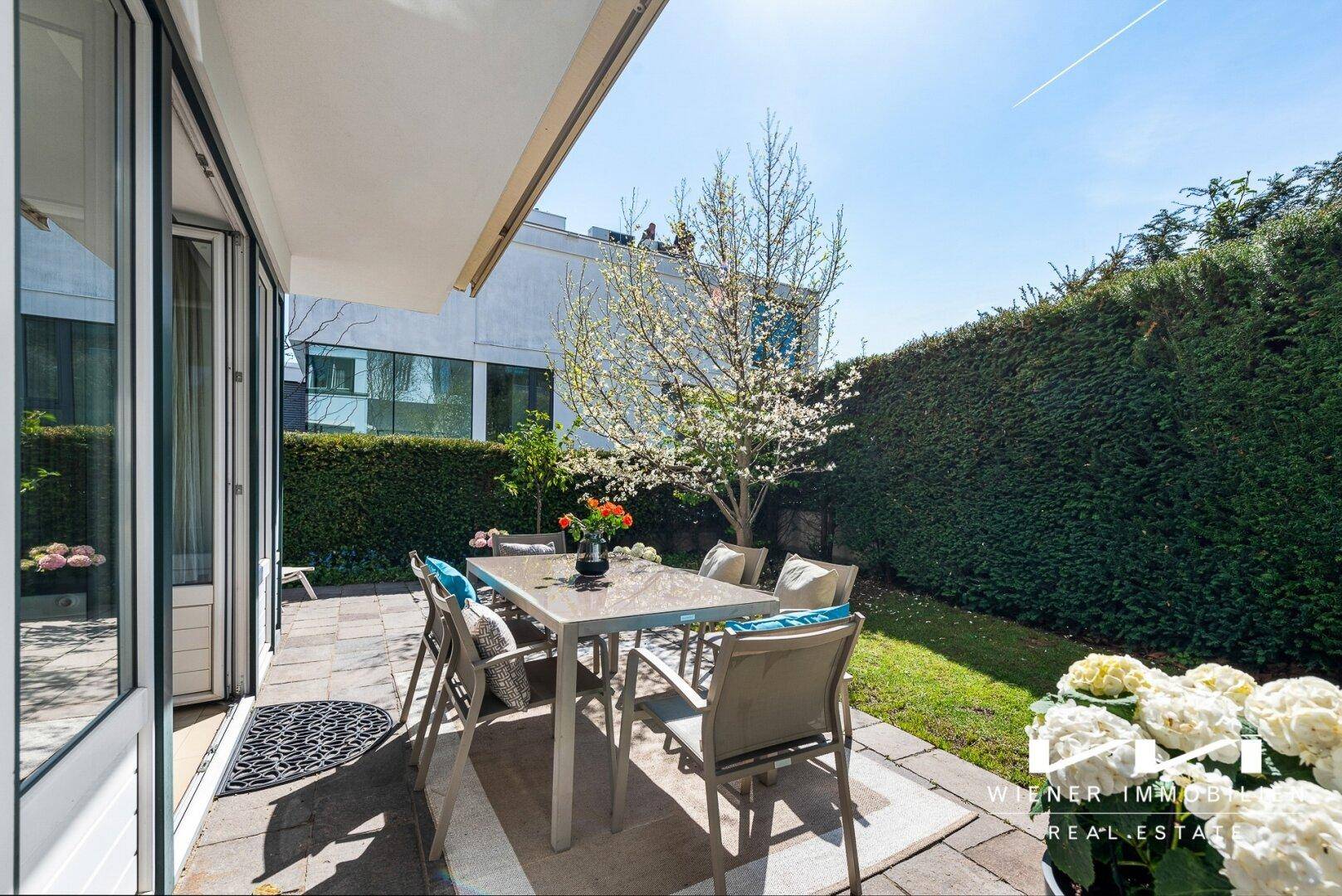 Helle Terrasse mit Esstisch und Stühlen, umgeben von Rasen und einer hohen Hecke.