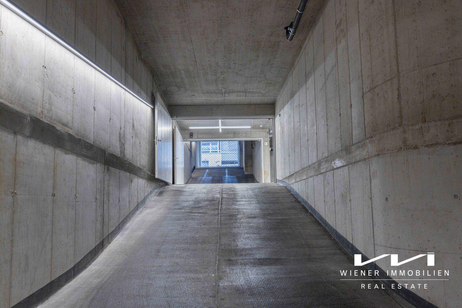 Abfahrt in eine Tiefgarage mit Betonwänden und Decken, beleuchtet von modernen Leuchtstoffröhren.