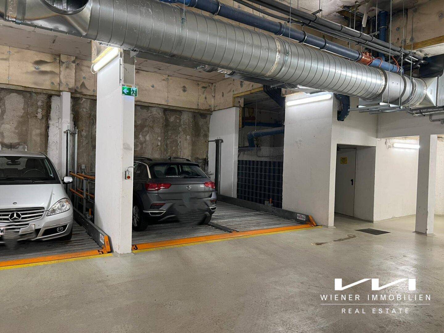 Übersicht der Tiefgarage mit mehreren mechanischen Parkplätzen und sichtbaren Lüftungsrohren an der Decke.