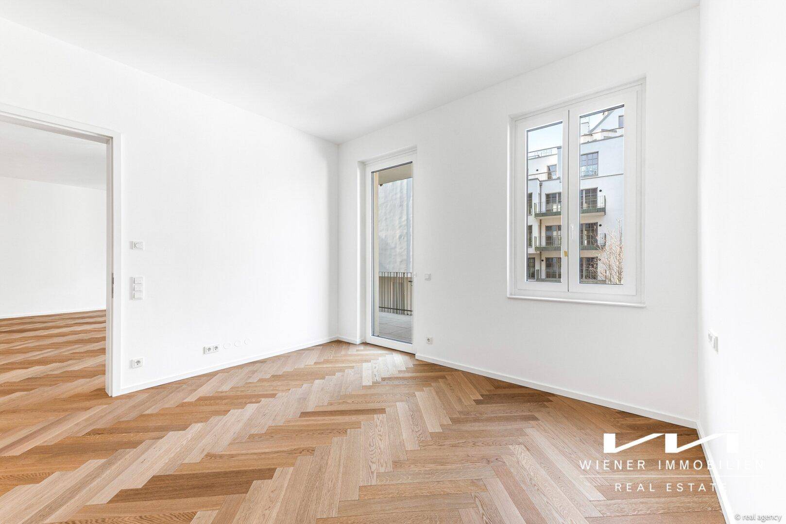 Freundliches Zimmer mit Fischgrätparkett, weißen Wänden und Zugang zu einem Balkon oder einer kleinen Terrasse.