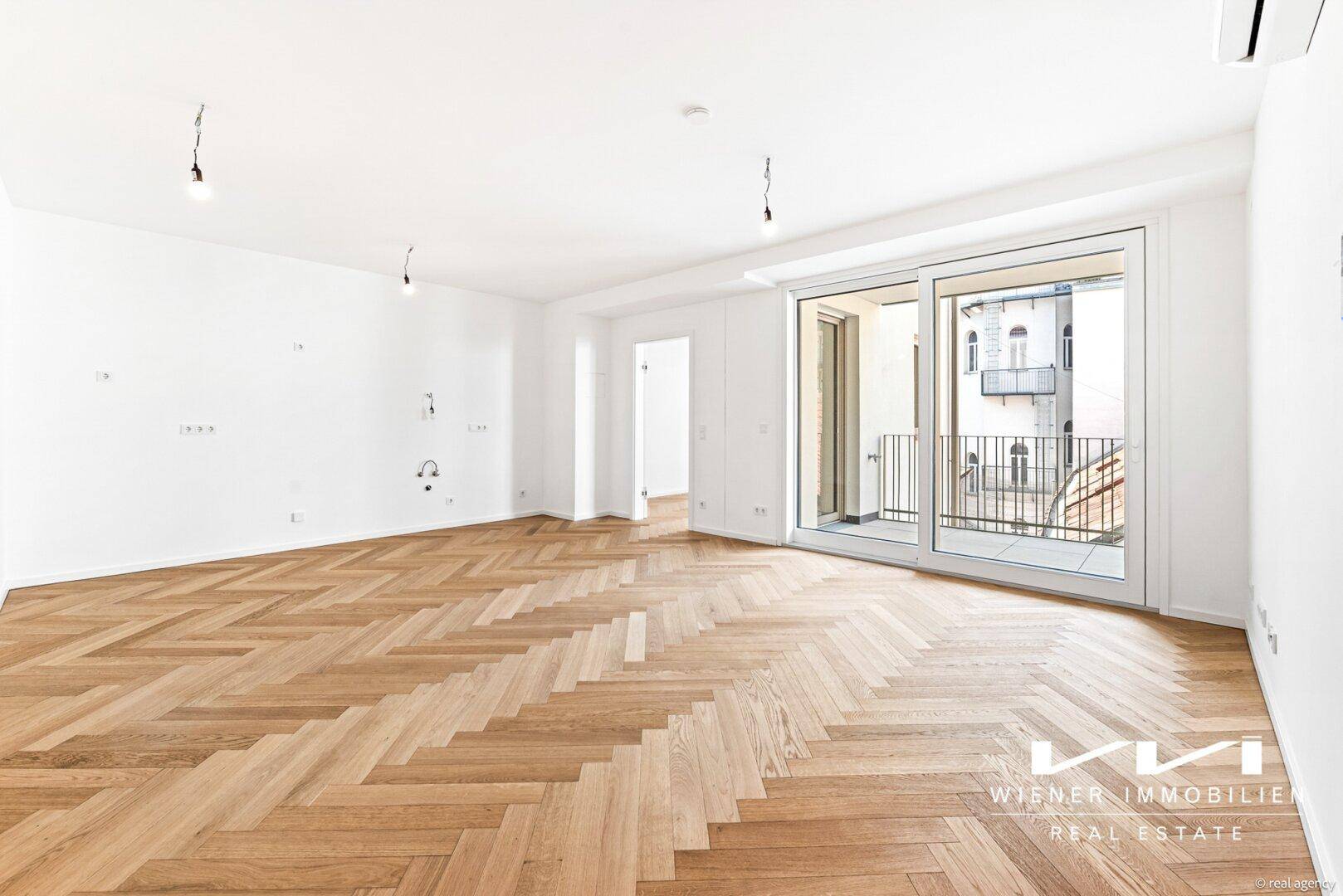 Heller Wohnbereich mit Fischgrätparkett, weißen Wänden und Zugang zu einem französischen Balkon.