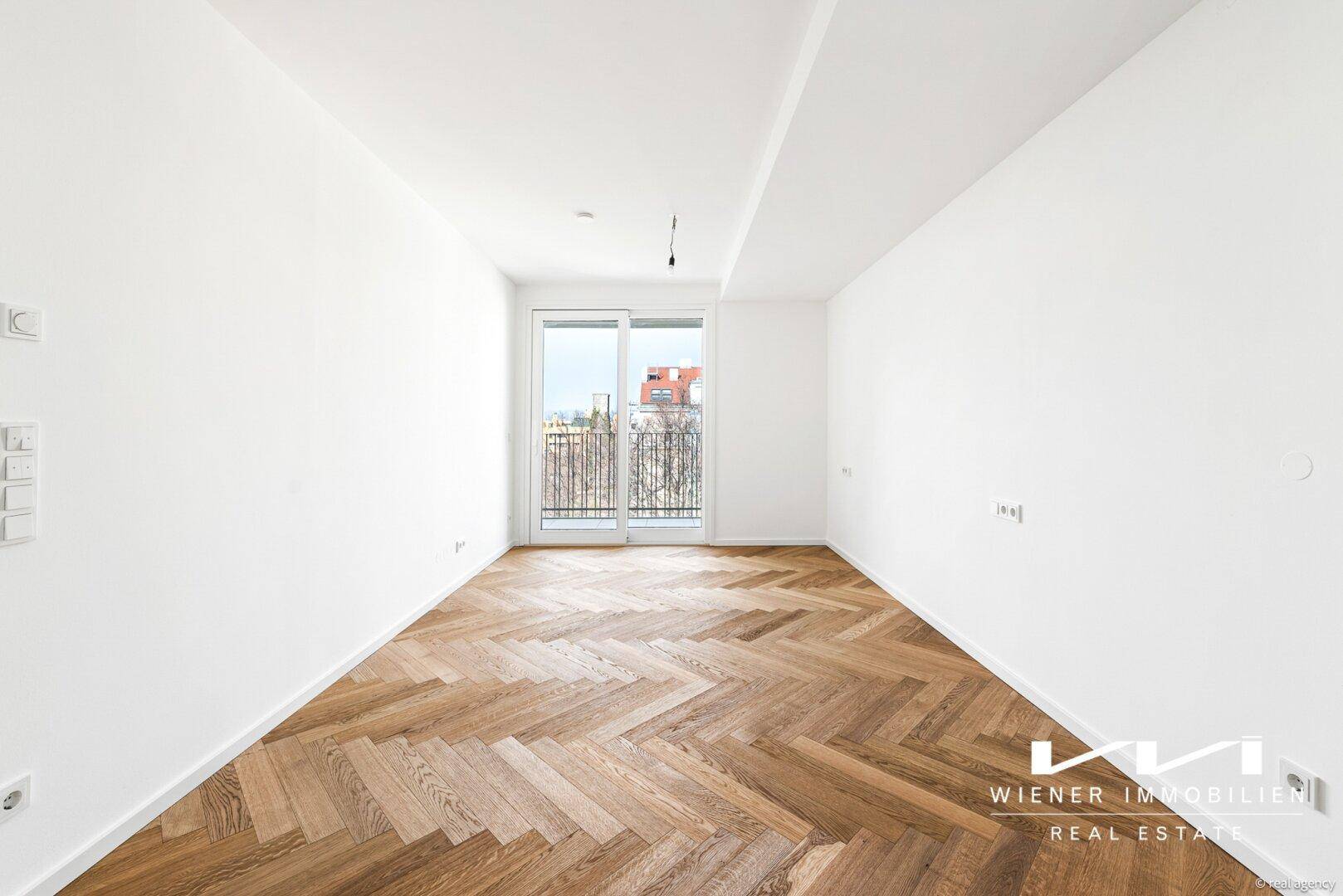 Helles, leeres Zimmer mit Fischgrätparkettboden und Zugang zu einem kleinen französischen Balkon.