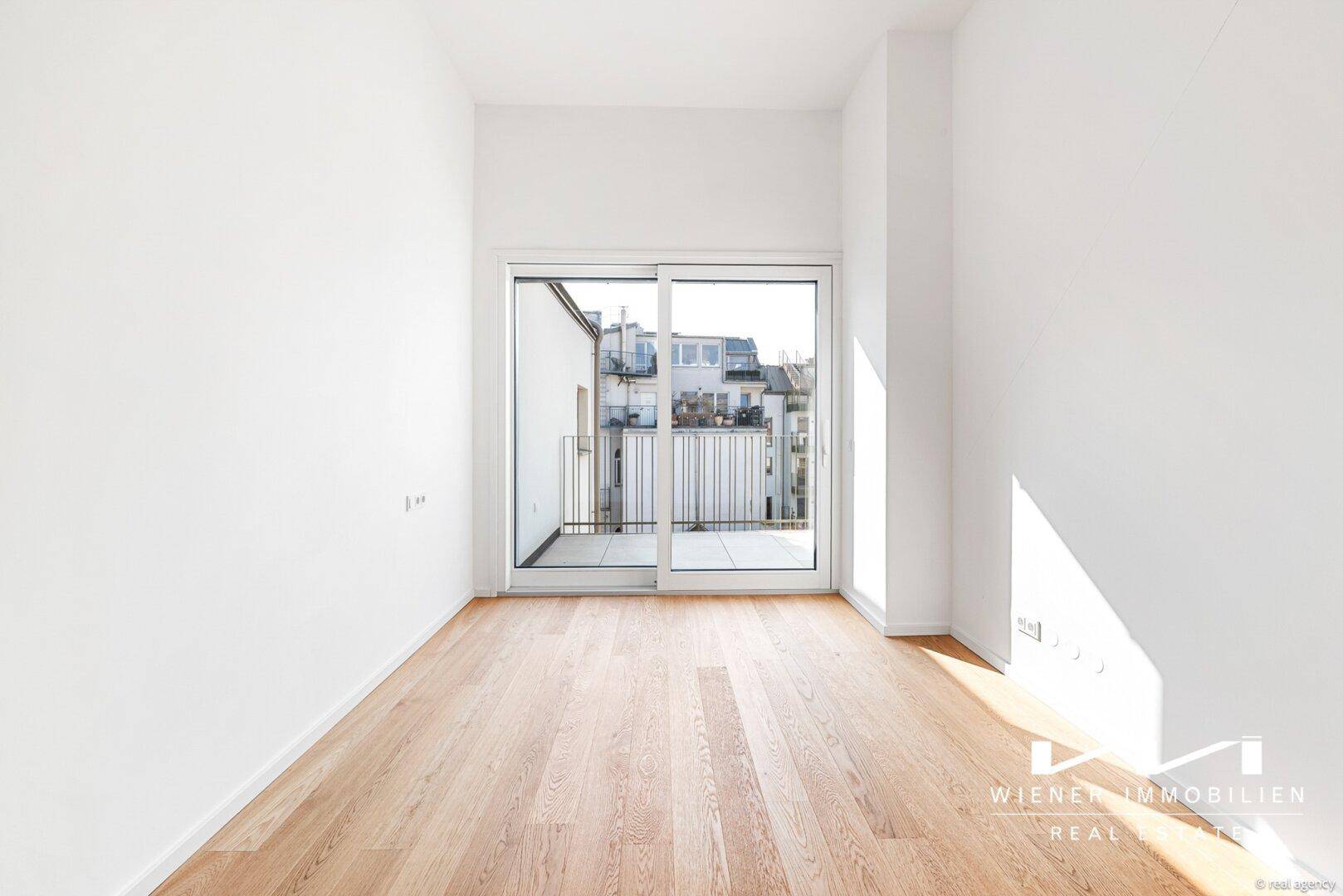 Helles Zimmer mit Holzboden und großer Schiebetür, die Zugang zu einem kleinen Balkon bietet.