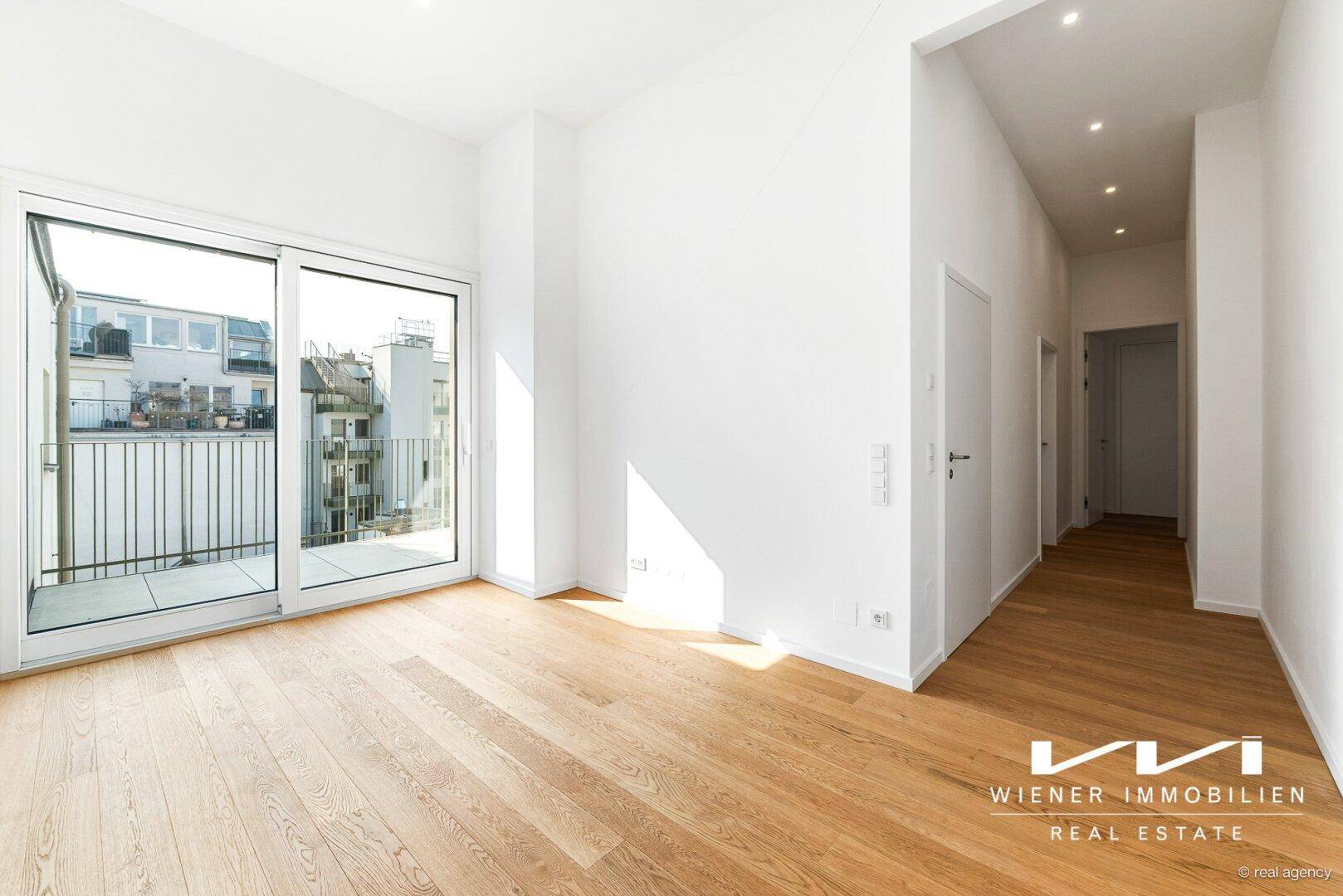 Heller Raum mit Holzboden und großzügiger Fensterfront, die den Zugang zu einem Balkon ermöglicht.