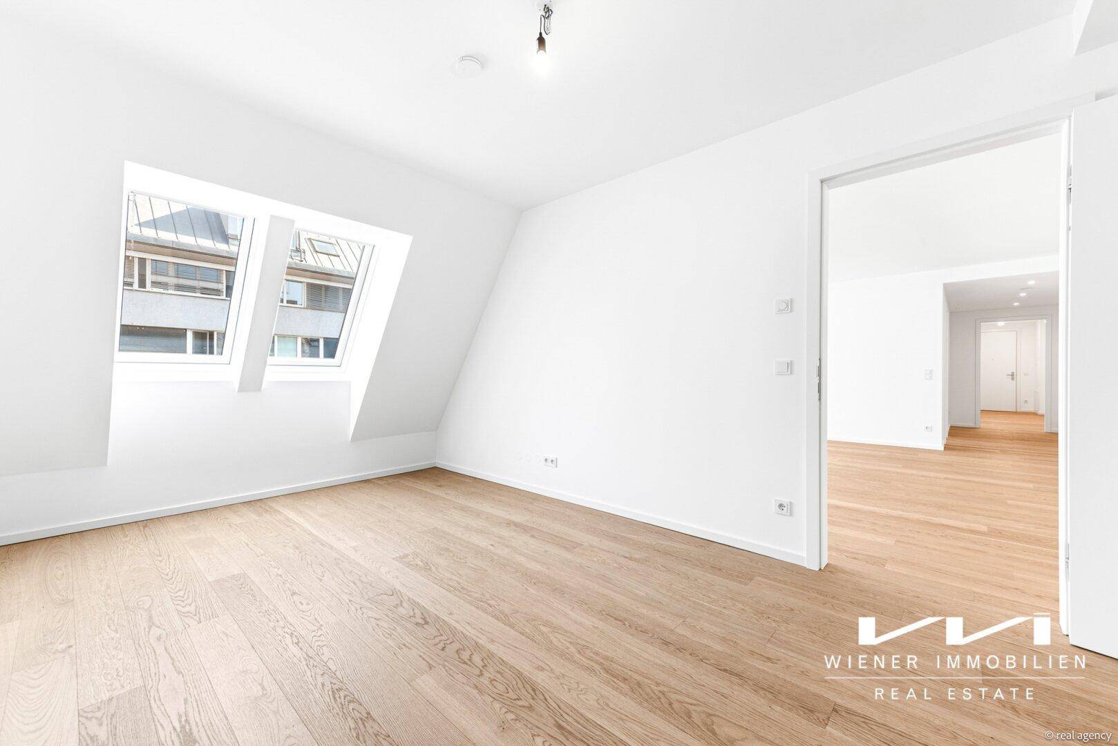 Heller Wohnraum mit Holzboden und Dachschrägenfenstern, der zu einem weiteren Zimmer führt.