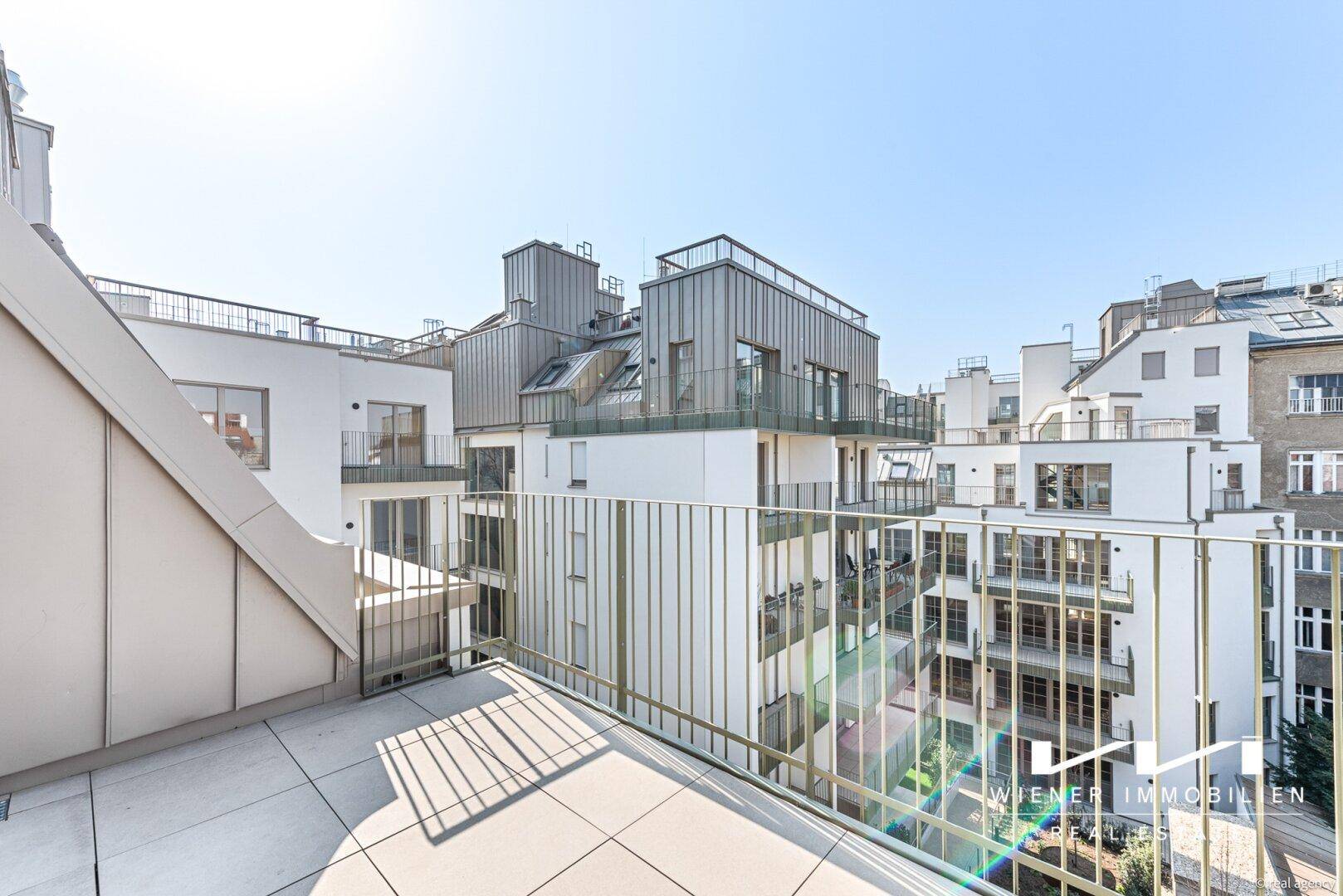 Geräumiger Balkon mit Fliesenboden und Blick auf moderne Dachlandschaft und umliegende Gebäude.