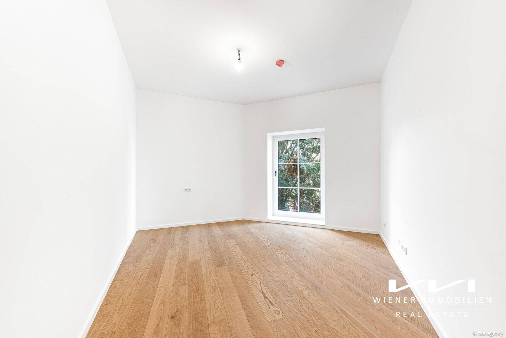 Kleines, helles Zimmer mit Parkettboden und Fenster mit Blick ins Grüne.