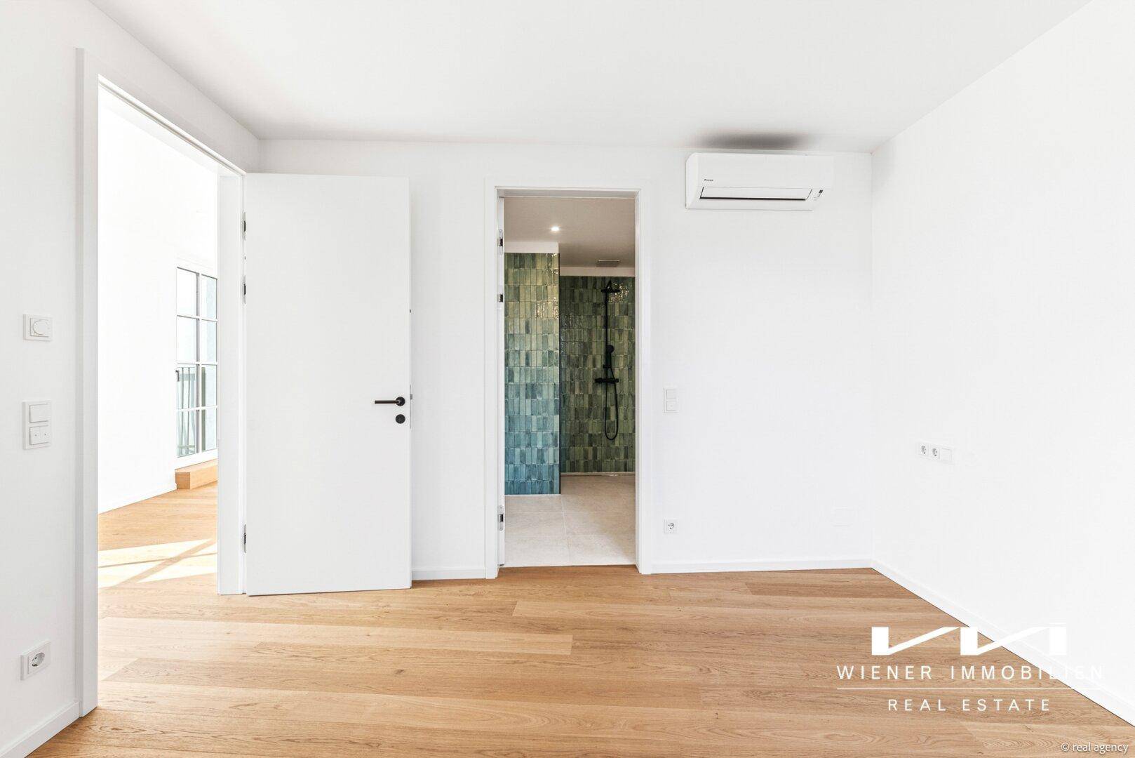 Helles Schlafzimmer mit Holzparkett und Zugang zu einem modernen Badezimmer mit grünen Fliesen.