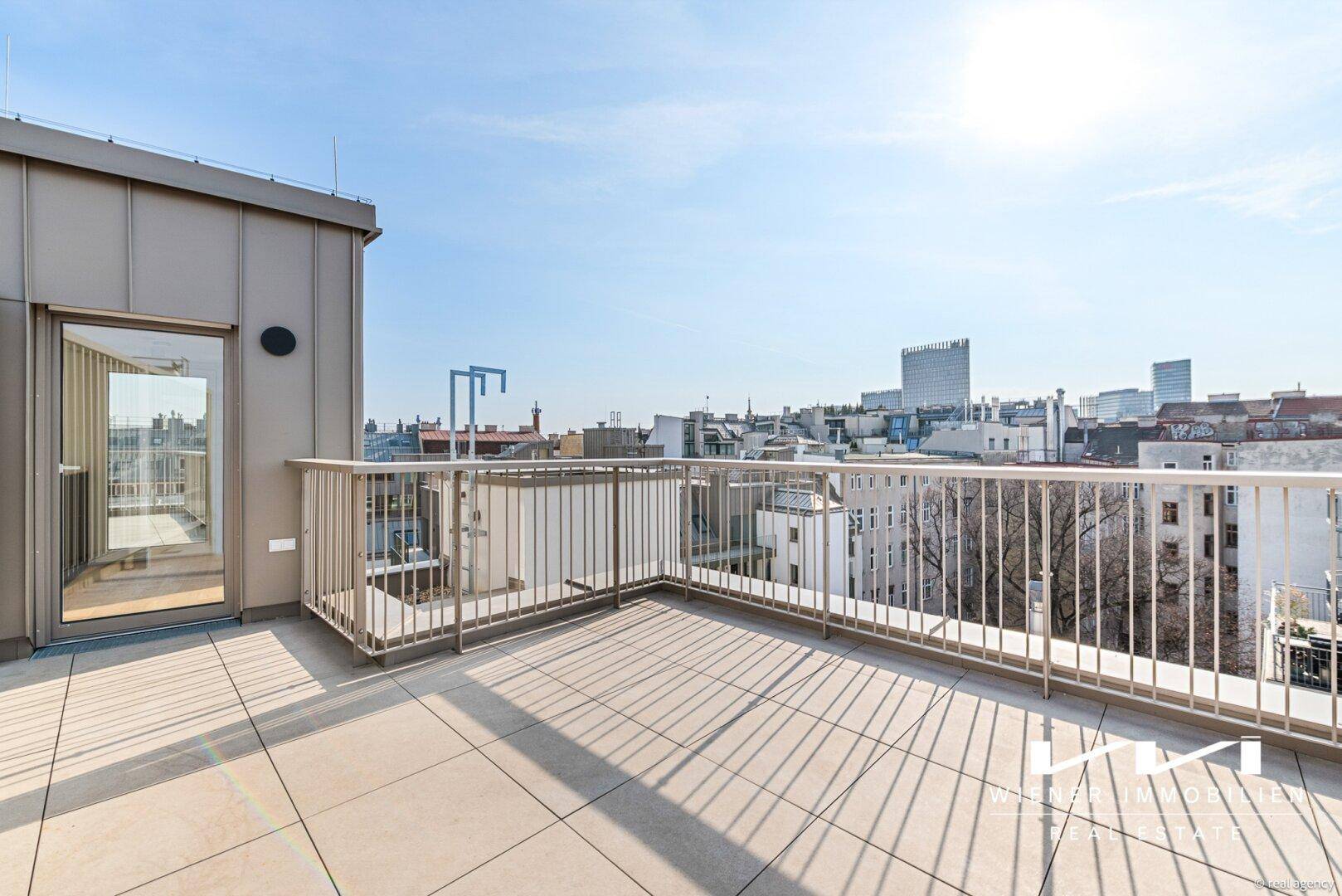 Geräumige Dachterrasse mit Fliesenboden und weitem Blick über die Stadt unter blauem Himmel.