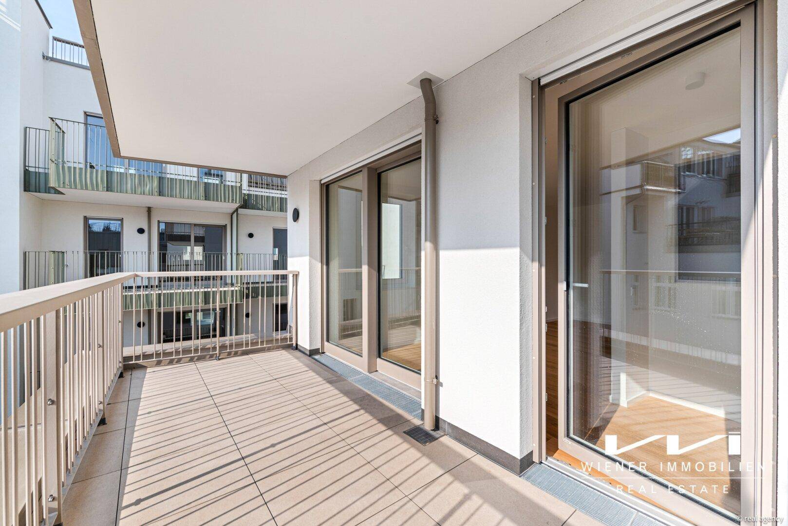 Heller Balkon mit Fliesenboden und Blick auf den Innenhof, zugänglich durch große Glasschiebetüren.