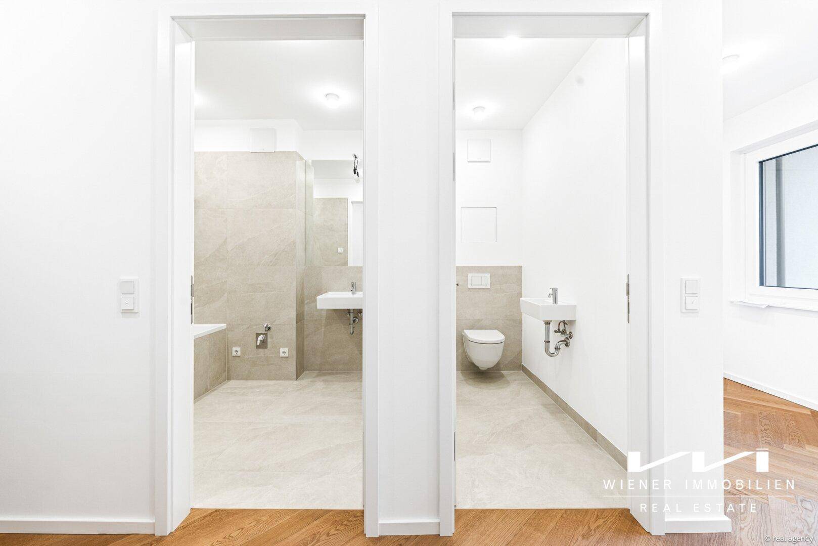 Blick vom Flur in ein modernes Badezimmer und eine separate Gästetoilette mit hellen Fliesen.