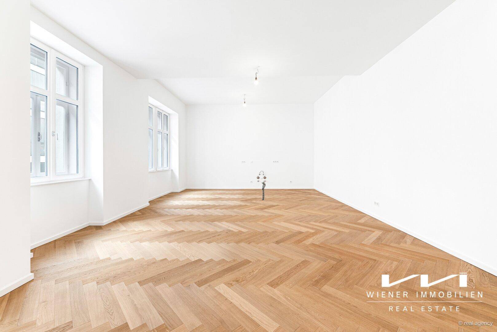 Großer, lichtdurchfluteter Raum mit elegantem Fischgrätparkett und hohen Decken.
