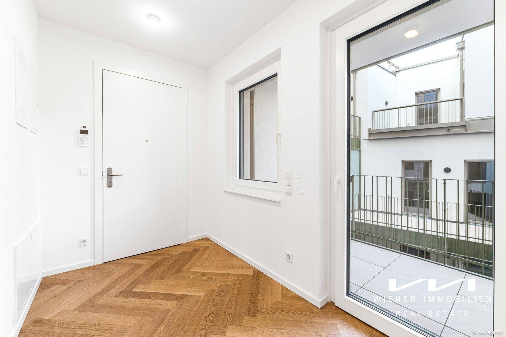 Eingangsbereich mit Fischgrätparkett und großer Glastür, die zum Balkon führt und den Innenhof zeigt.