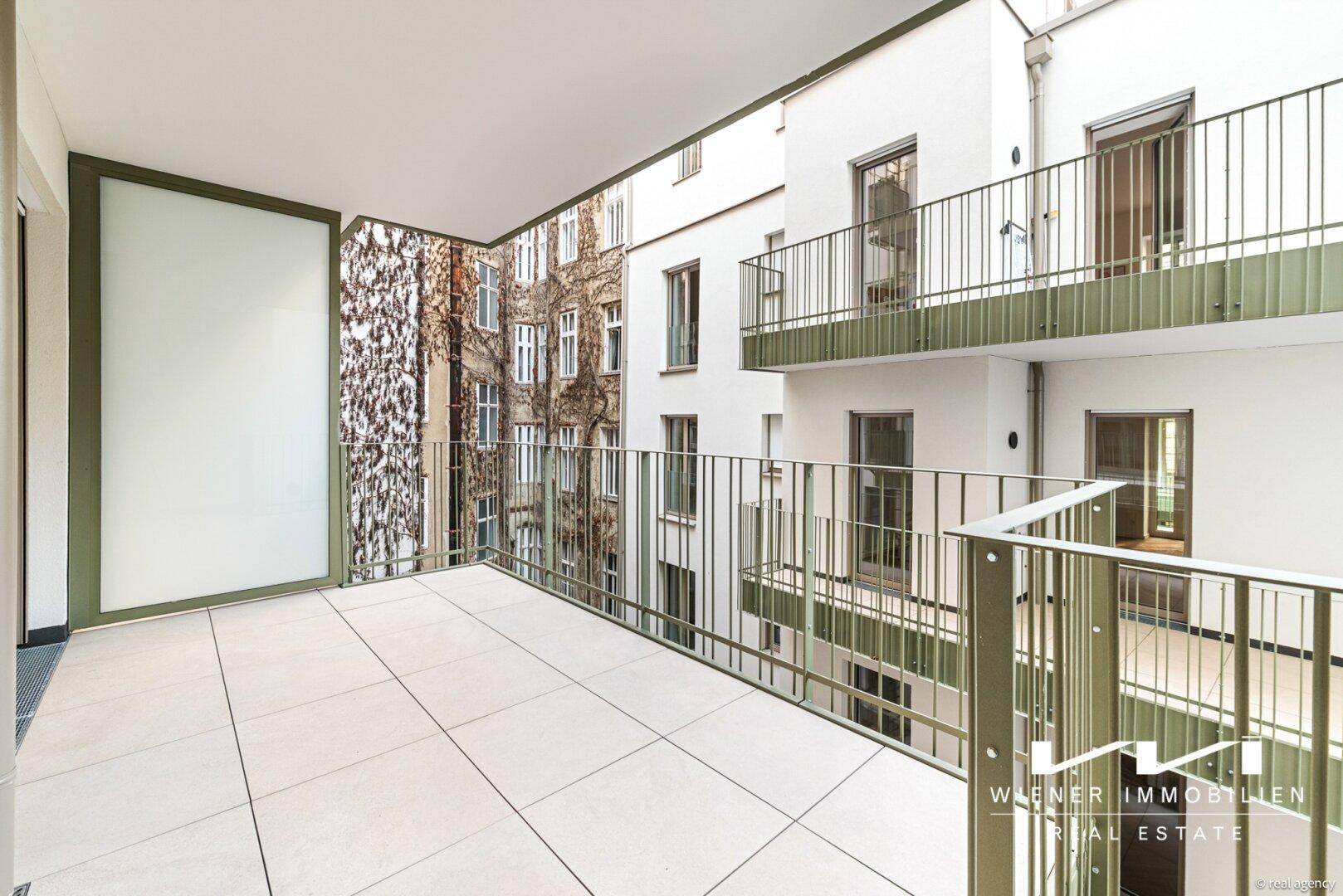 Heller Balkon mit Fliesenboden und Blick auf den begrünten Innenhof des Gebäudes.