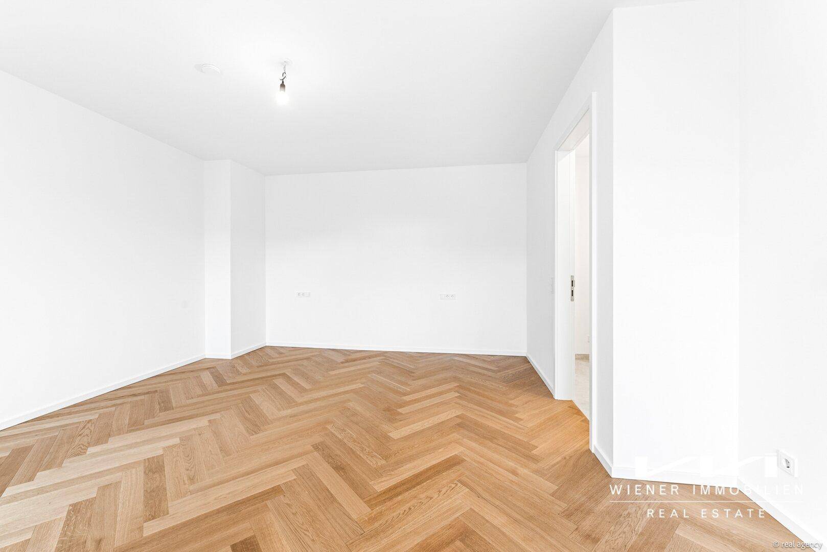 Geräumiges, helles Zimmer mit elegantem Fischgrätparkett und weißen Wänden.