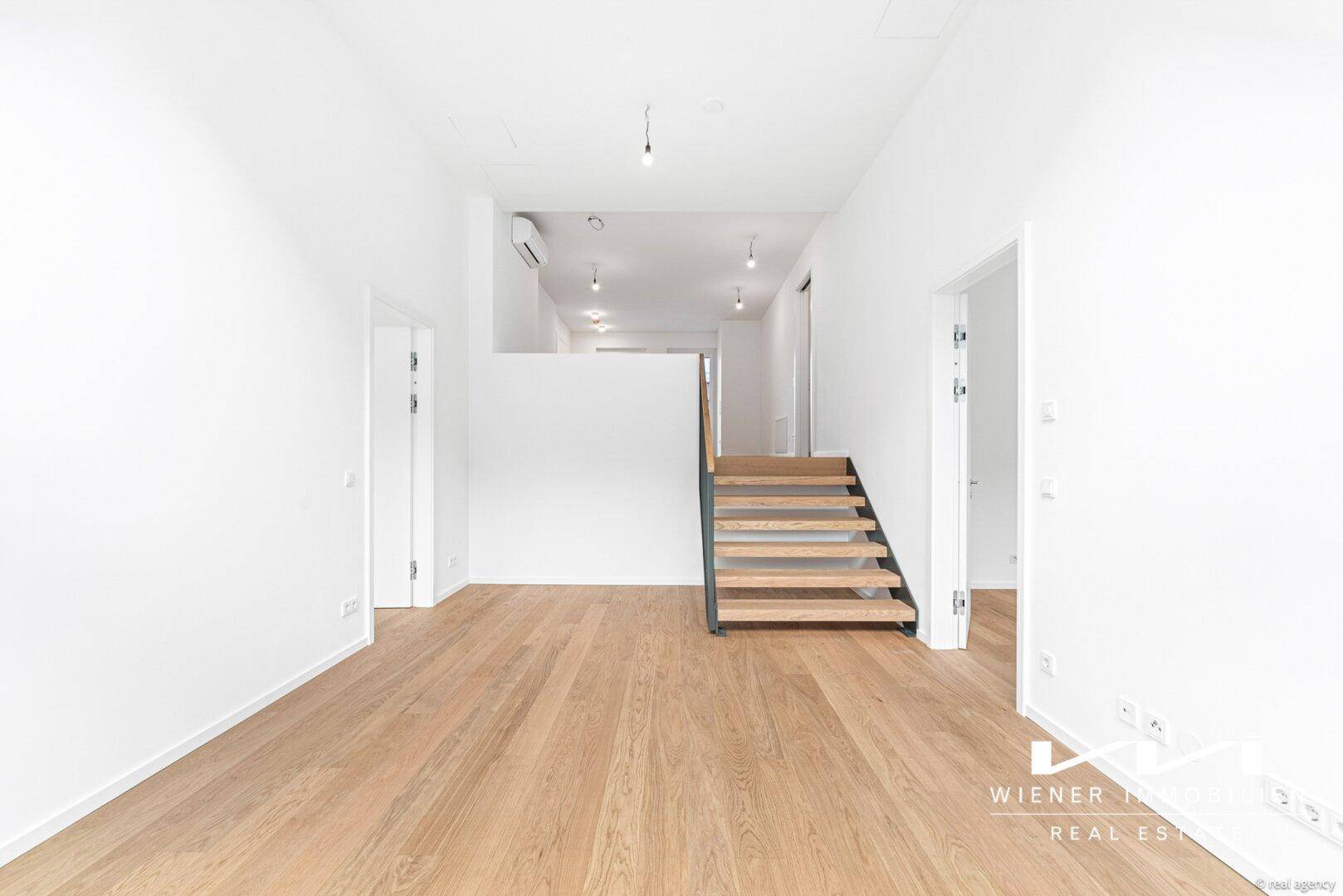 Offener Wohnbereich mit Holzfußboden, moderner Treppe und Zugang zu weiteren Räumen.