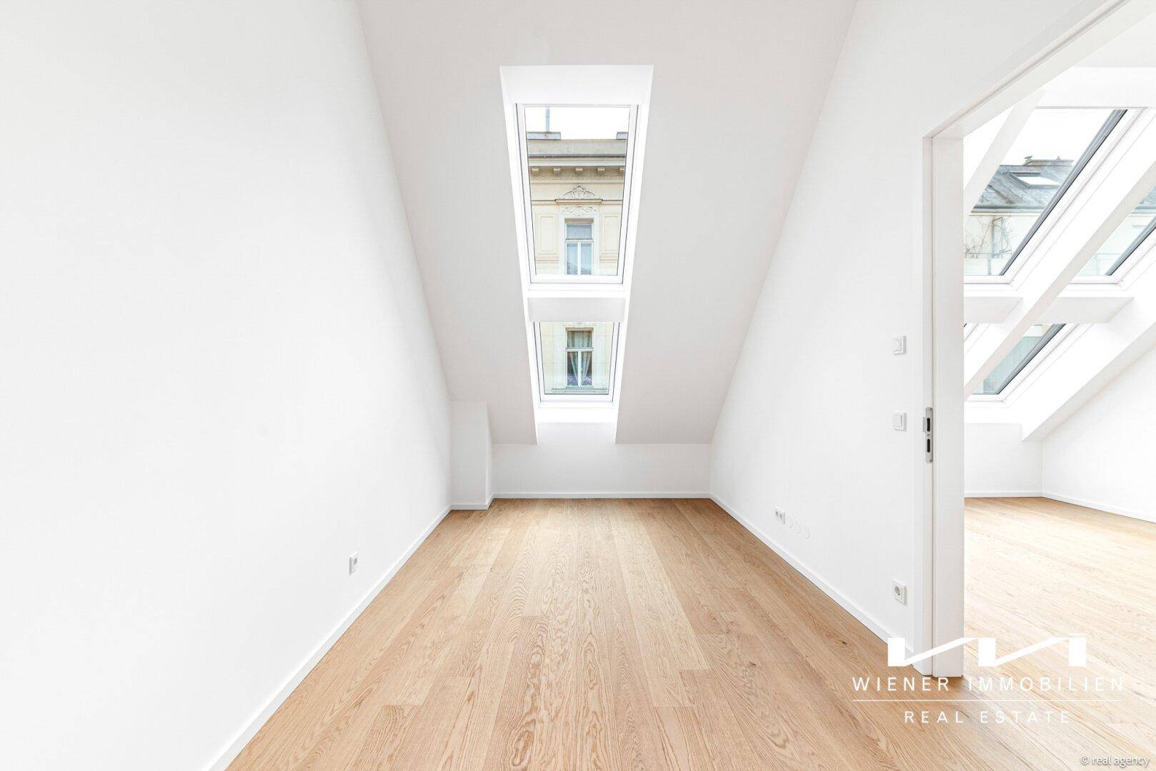 Dachgeschosszimmer mit schrägen Wänden, Holzfußboden und vertikalen Dachfenstern.