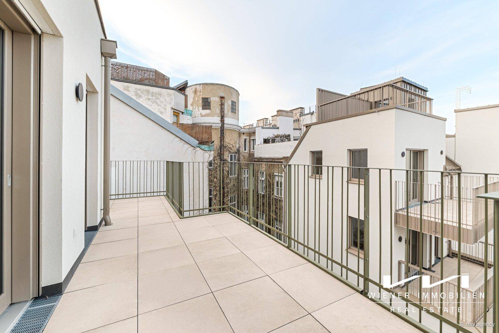 Langer Balkon mit Fliesenboden und Blick auf die umliegenden Gebäude und Dächer der Stadt.