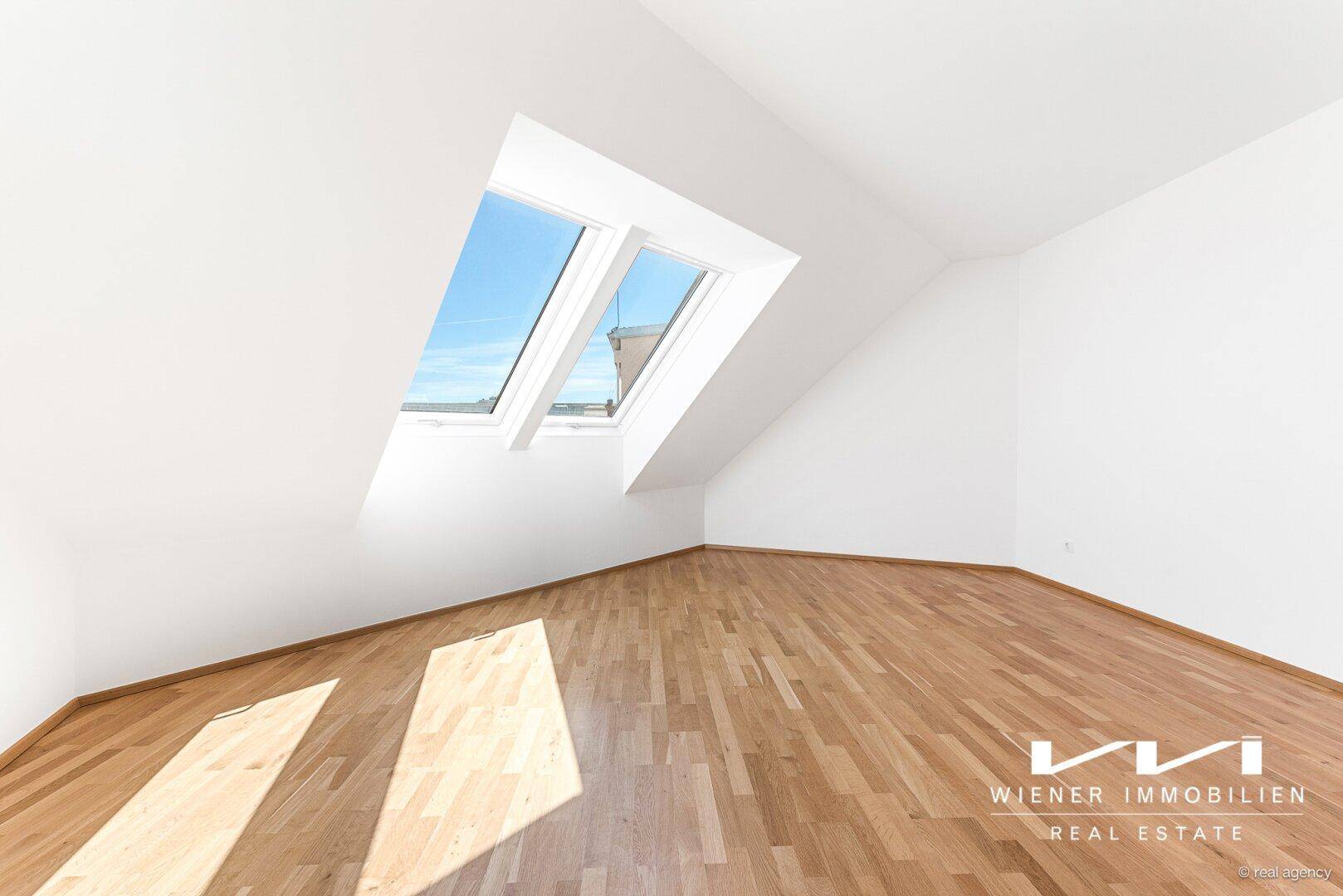Helles Zimmer mit Dachschrägen, Holzboden und zwei großen Dachfenstern, die viel Tageslicht hereinlassen.