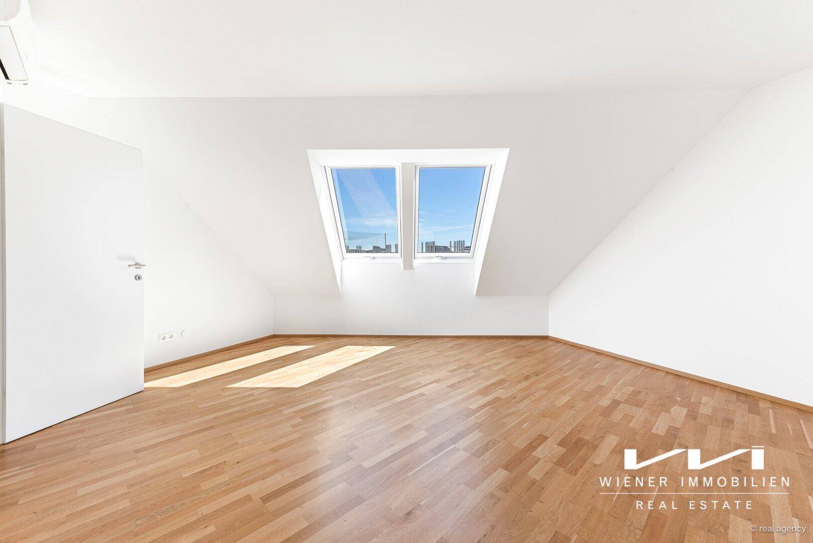 Geräumiges Zimmer mit hellen Holzböden und großen Dachfenstern, die den Raum mit Sonnenlicht durchfluten.
