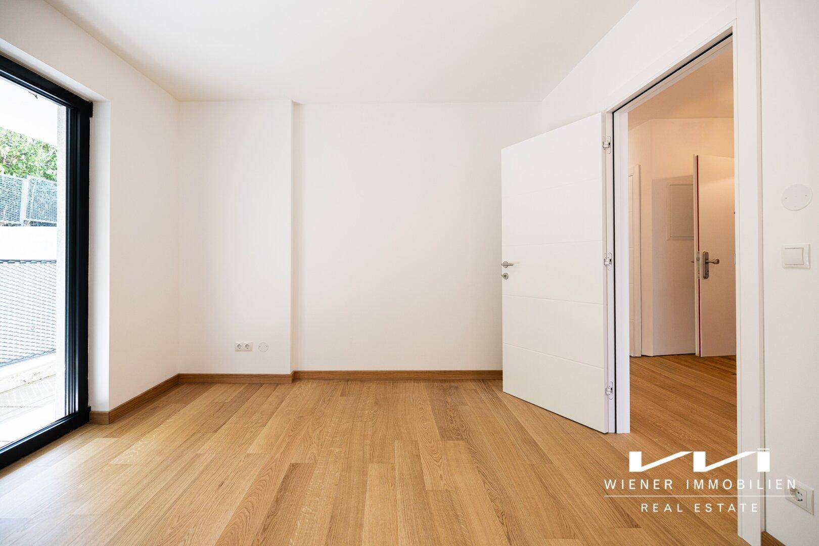 Geräumiges Zimmer mit hellem Parkettboden und offener Tür zu einem weiteren Raum.