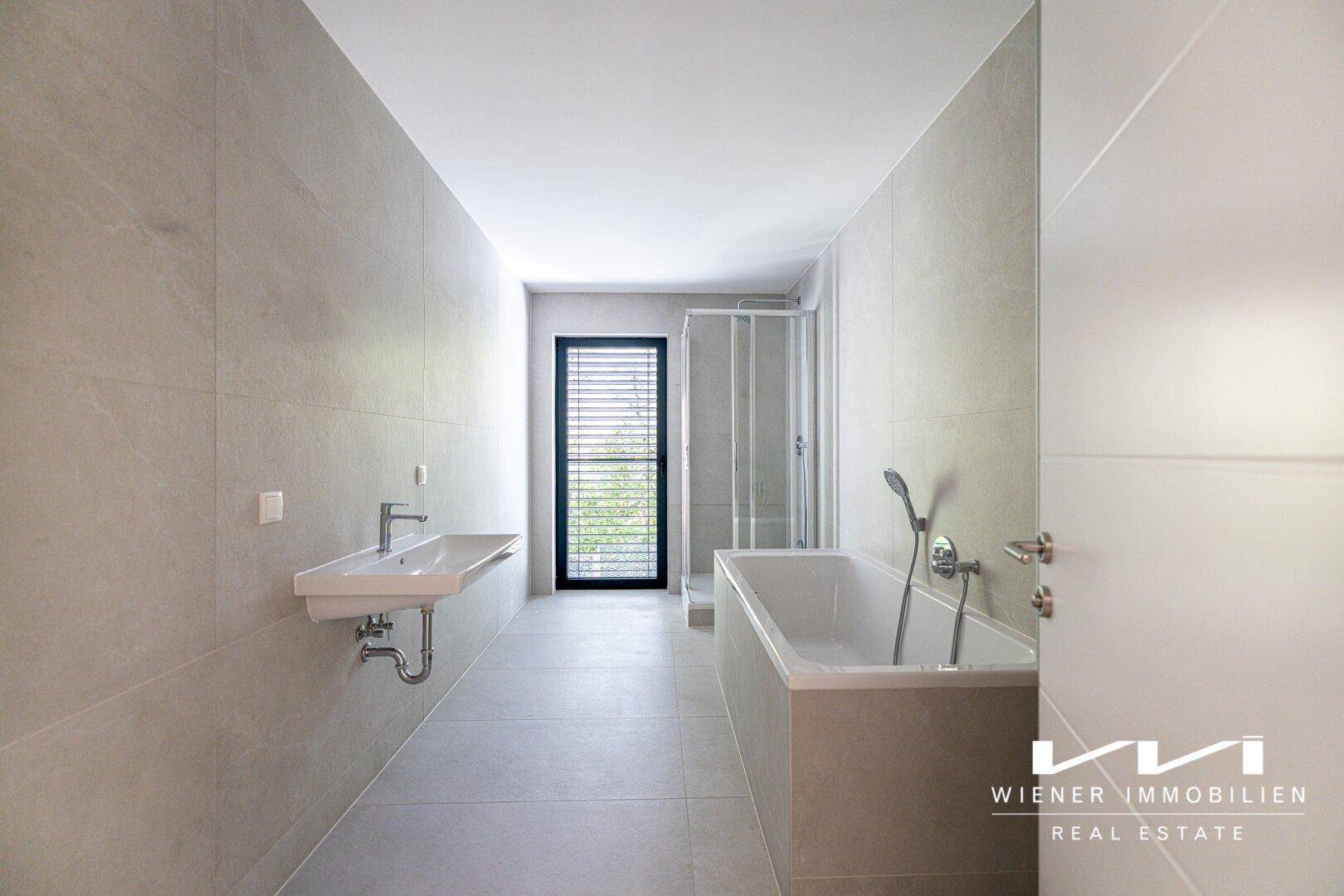 Modernes Badezimmer mit grauen Fliesen, Badewanne, Dusche und einem Fenster für Tageslicht.