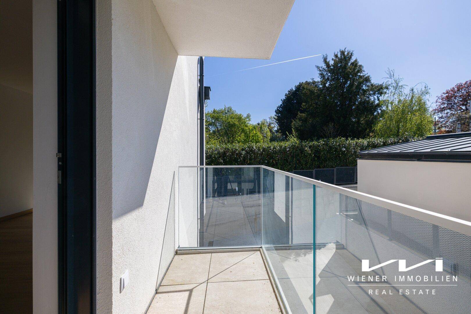 Sonniger Balkon mit Glasgeländer und Blick auf die grüne Umgebung und den blauen Himmel.