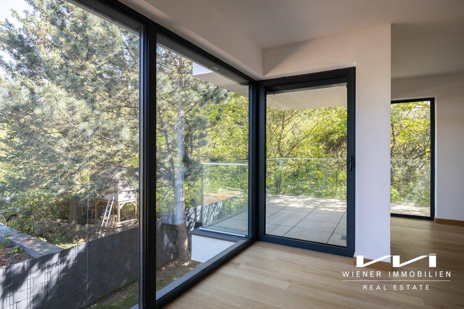 Heller Wohnraum mit großen Fenstern, Holzboden und Zugang zu einem Balkon mit Grünblick.