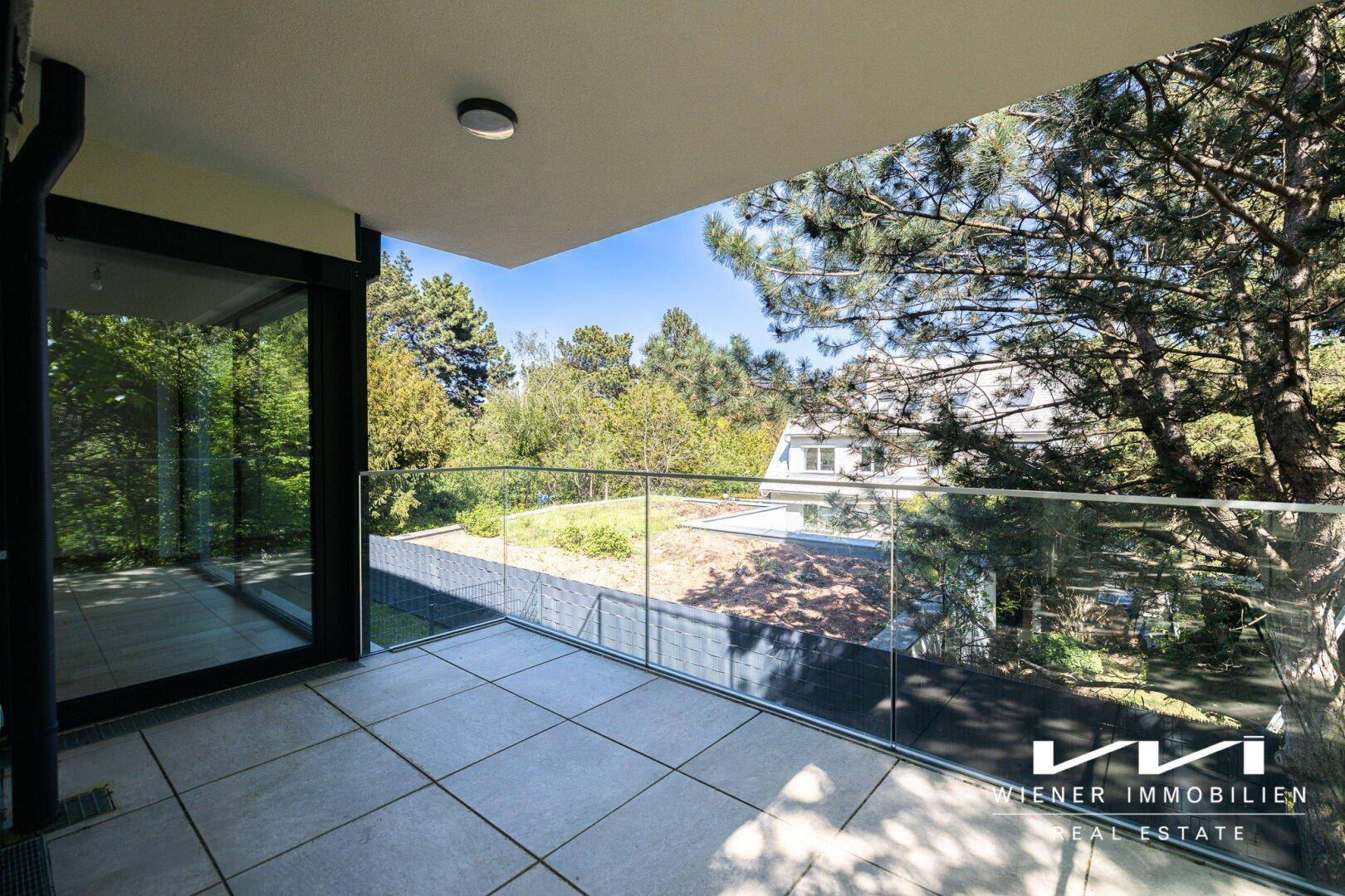 Großer Balkon mit Fliesenboden und Glasgeländer, umgeben von Bäumen und Grünflächen.