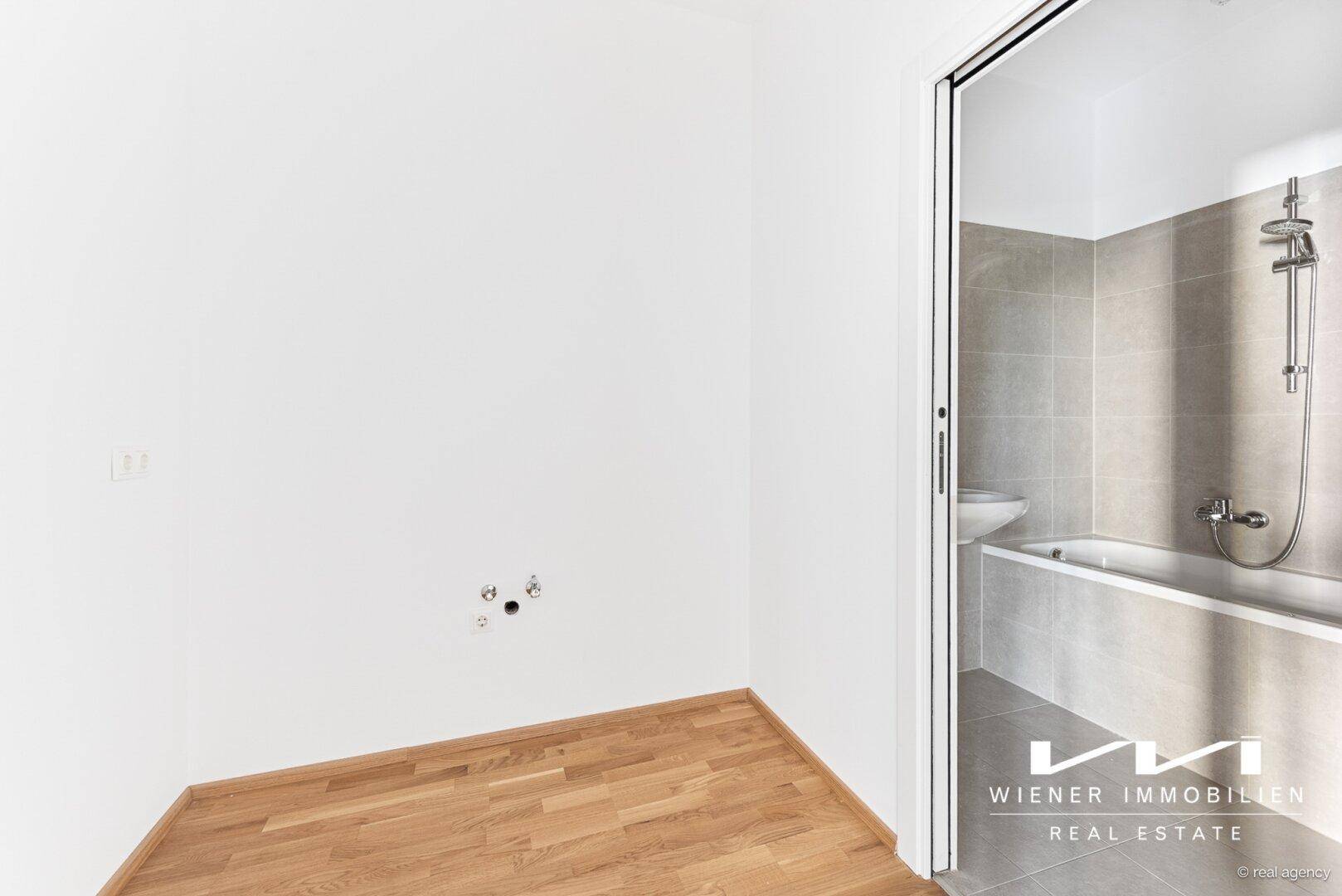Praktischer Raum mit Holzboden und Anschlüssen für eine Waschmaschine, angrenzend an ein Badezimmer.