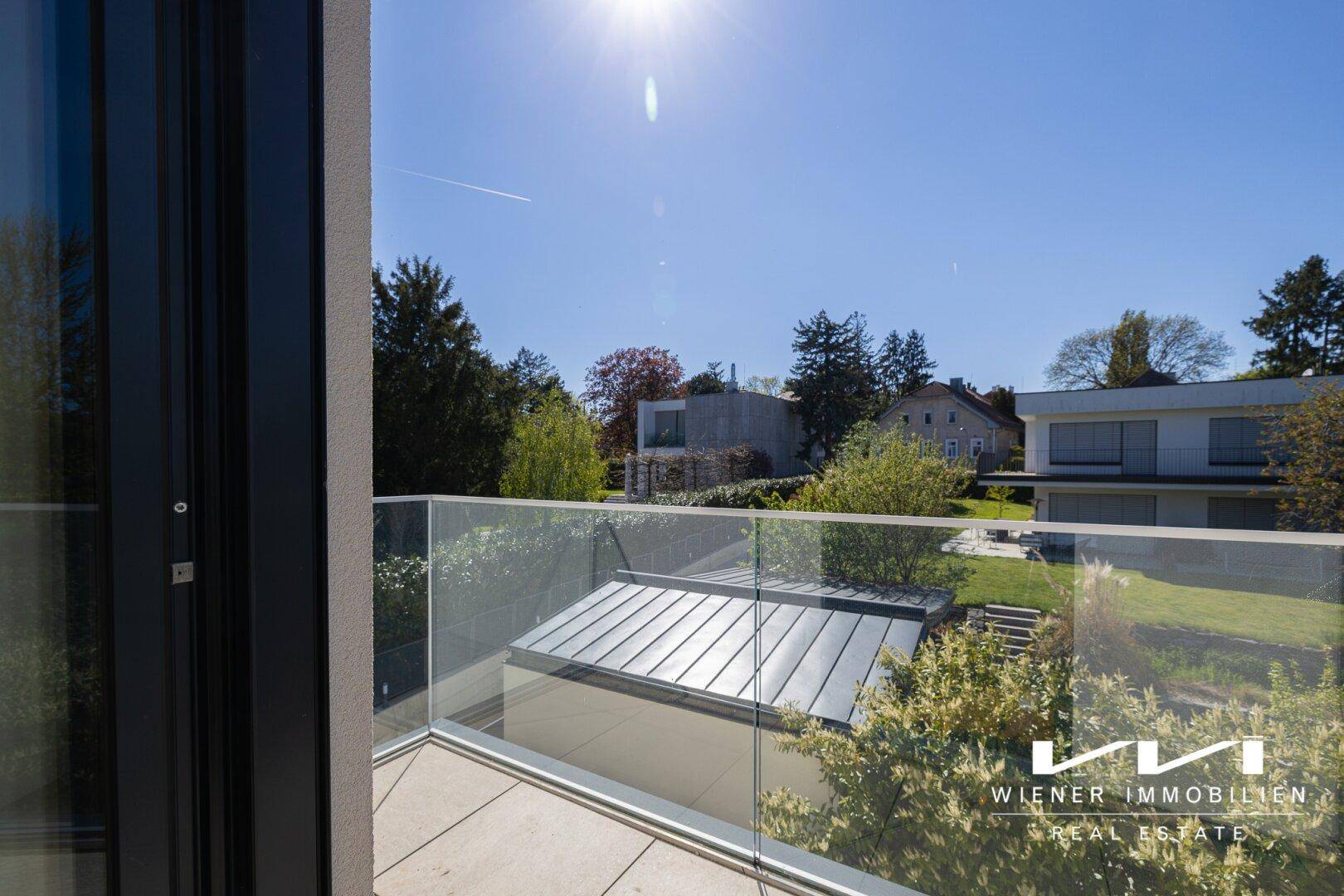 Heller Balkon mit modernem Glasgeländer und Blick auf benachbarte Häuser und grüne Gärten.