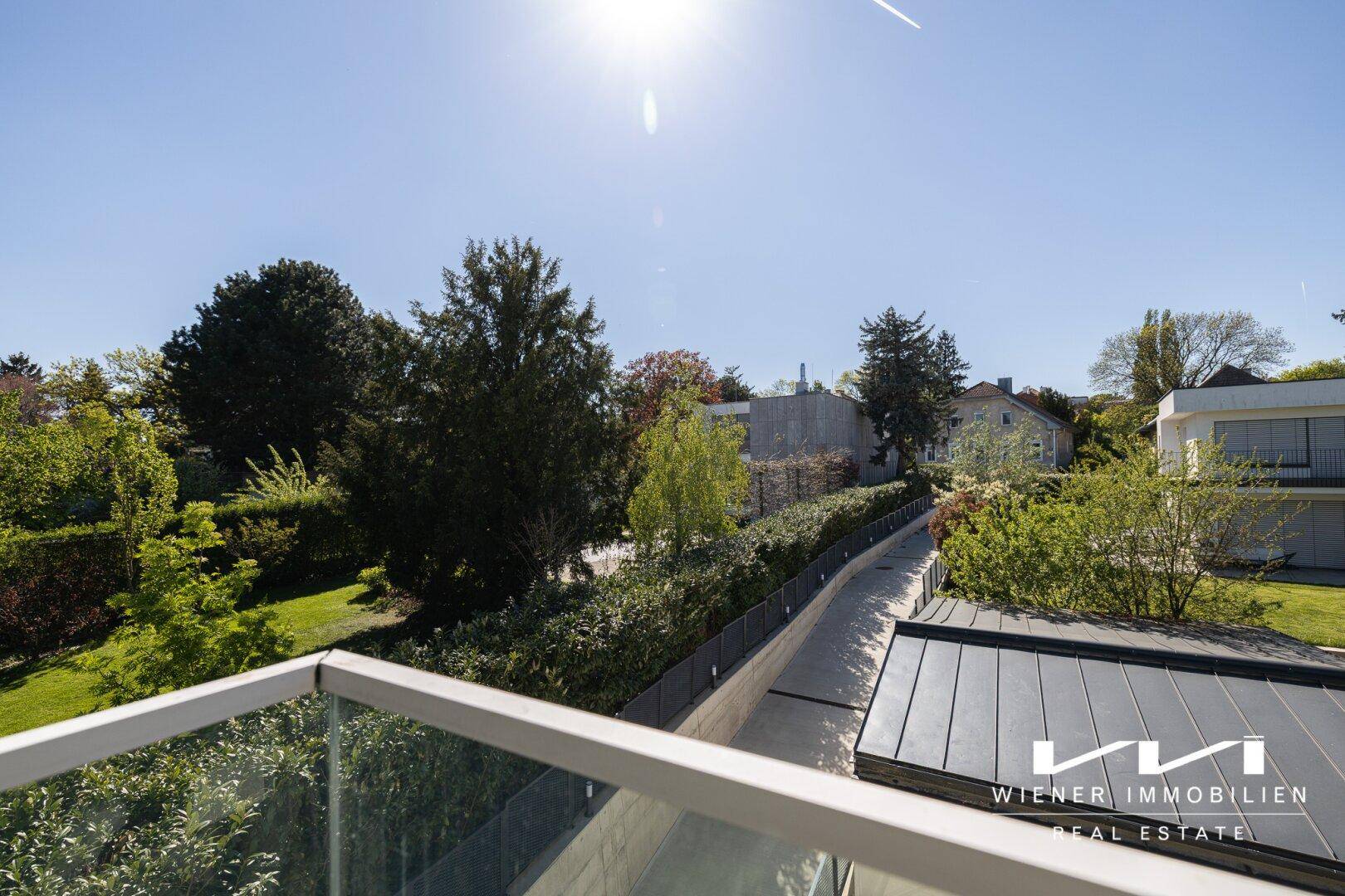Sonniger Balkon mit Glasgeländer, der einen Weg, Bäume und weitere Häuser in der Ferne zeigt.
