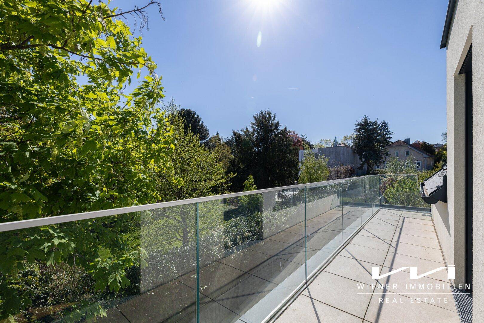 Sonniger Balkon mit Glasgeländer und Fliesenboden, umgeben von üppigem Grün und Bäumen.