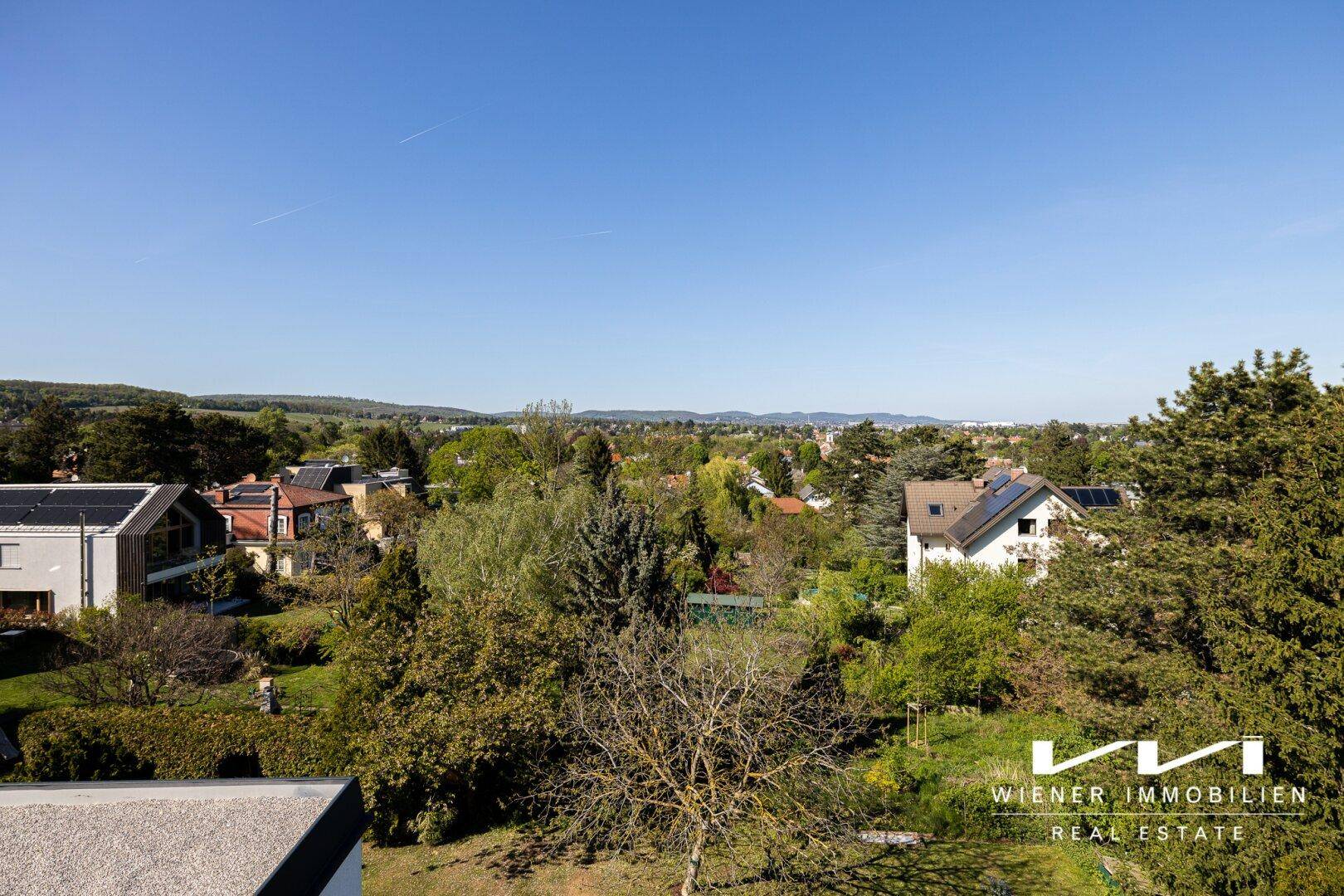 Weitläufiger Blick über eine grüne Wohngegend mit vielen Bäumen und Häusern unter blauem Himmel.