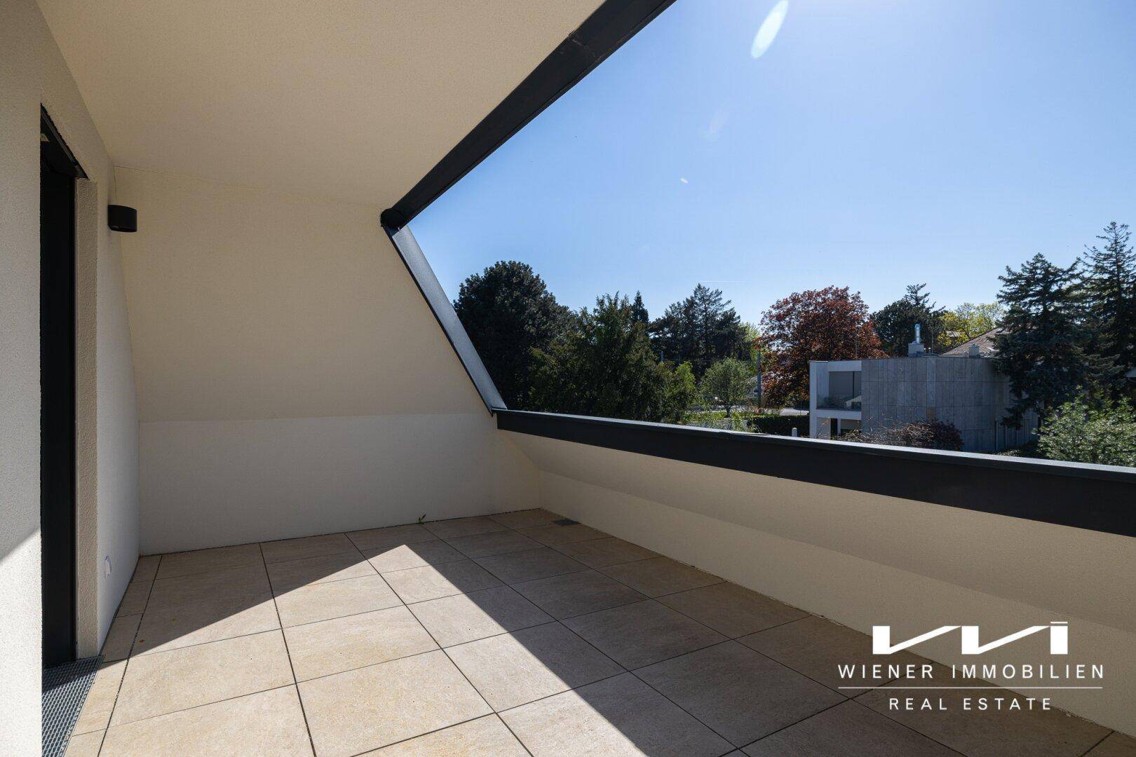 Sonniger Balkon mit Fliesenboden und schräger Dachkonstruktion, bietet Blick auf Bäume und Häuser.