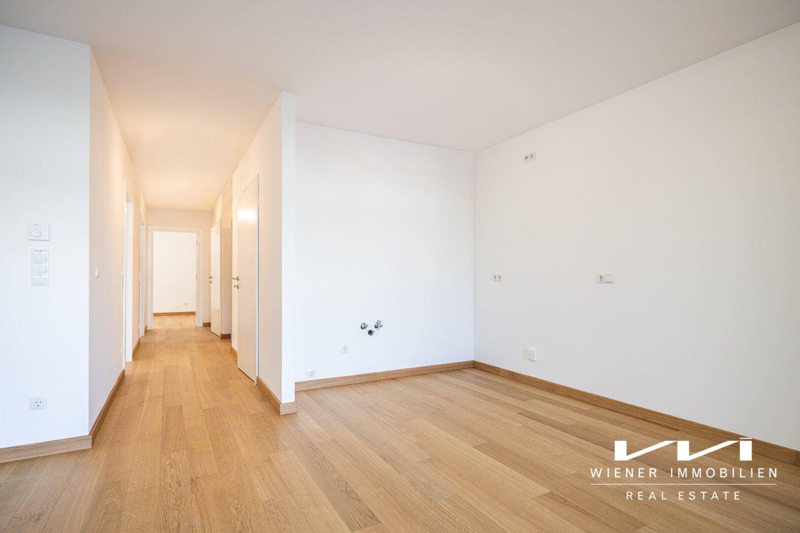 Heller Innenraum mit Holzboden und vorbereiteten Anschlüssen, möglicherweise für eine Küche oder ein Bad.