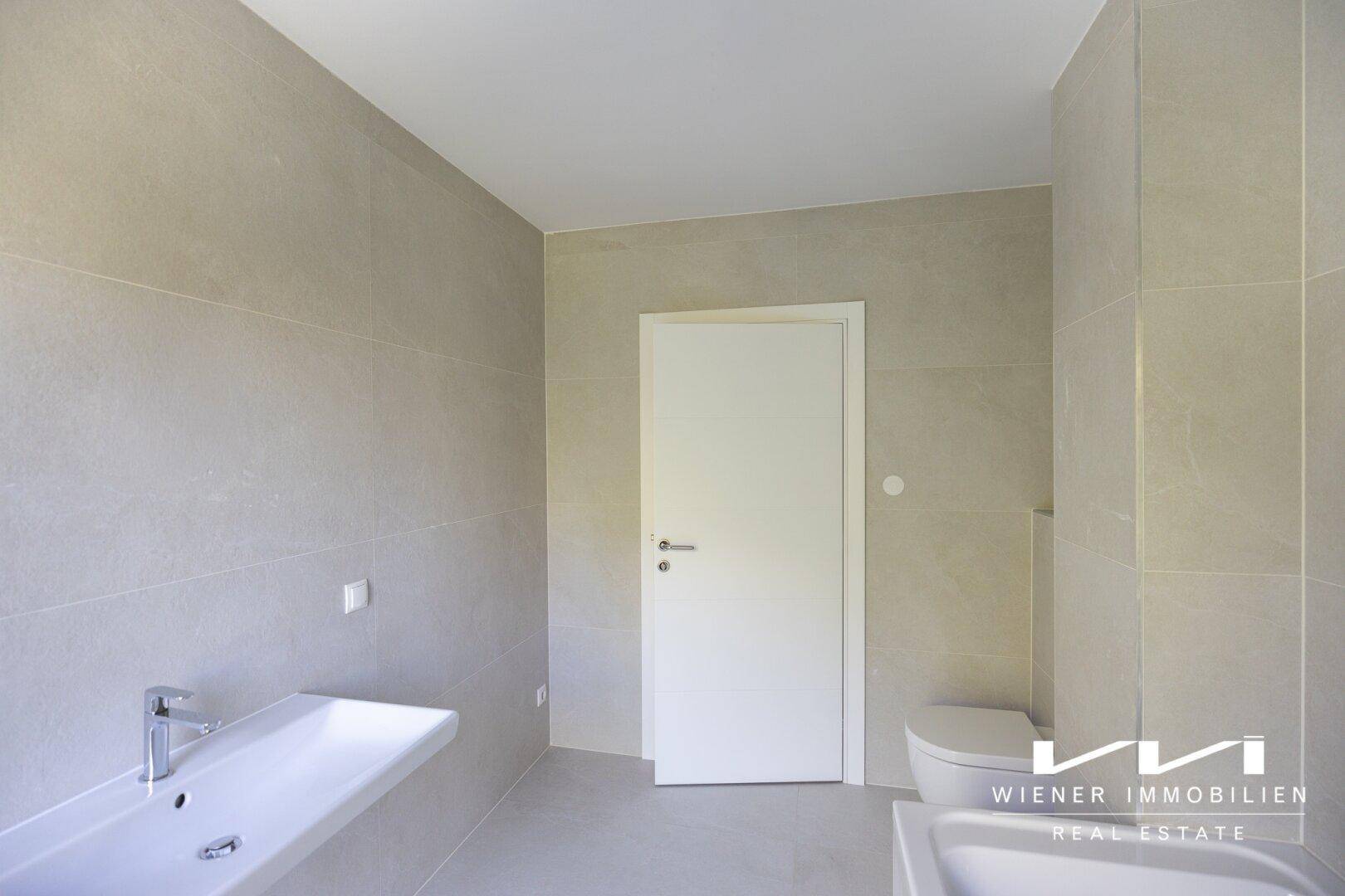 Modernes Badezimmer mit hellgrauen Fliesen, wandmontiertem Waschbecken und Badewanne.