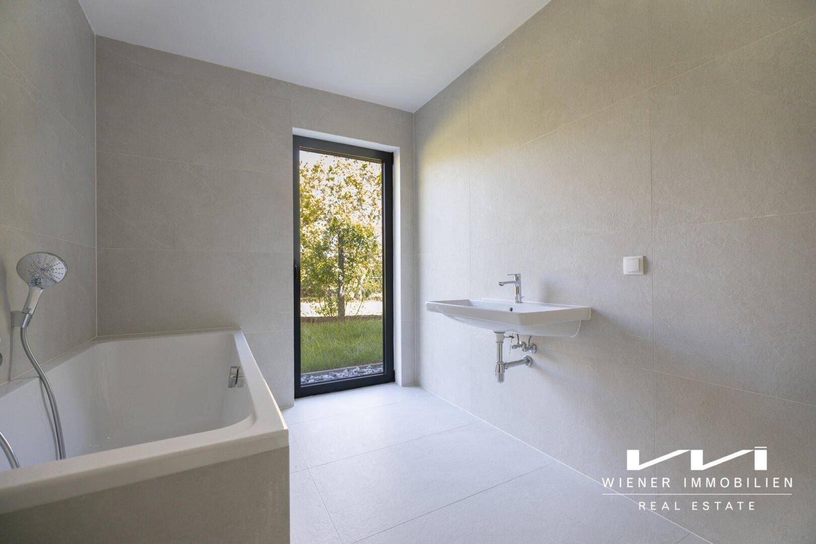 Helles Badezimmer mit Badewanne, wandmontiertem Waschbecken und großem Fenster zum Garten.
