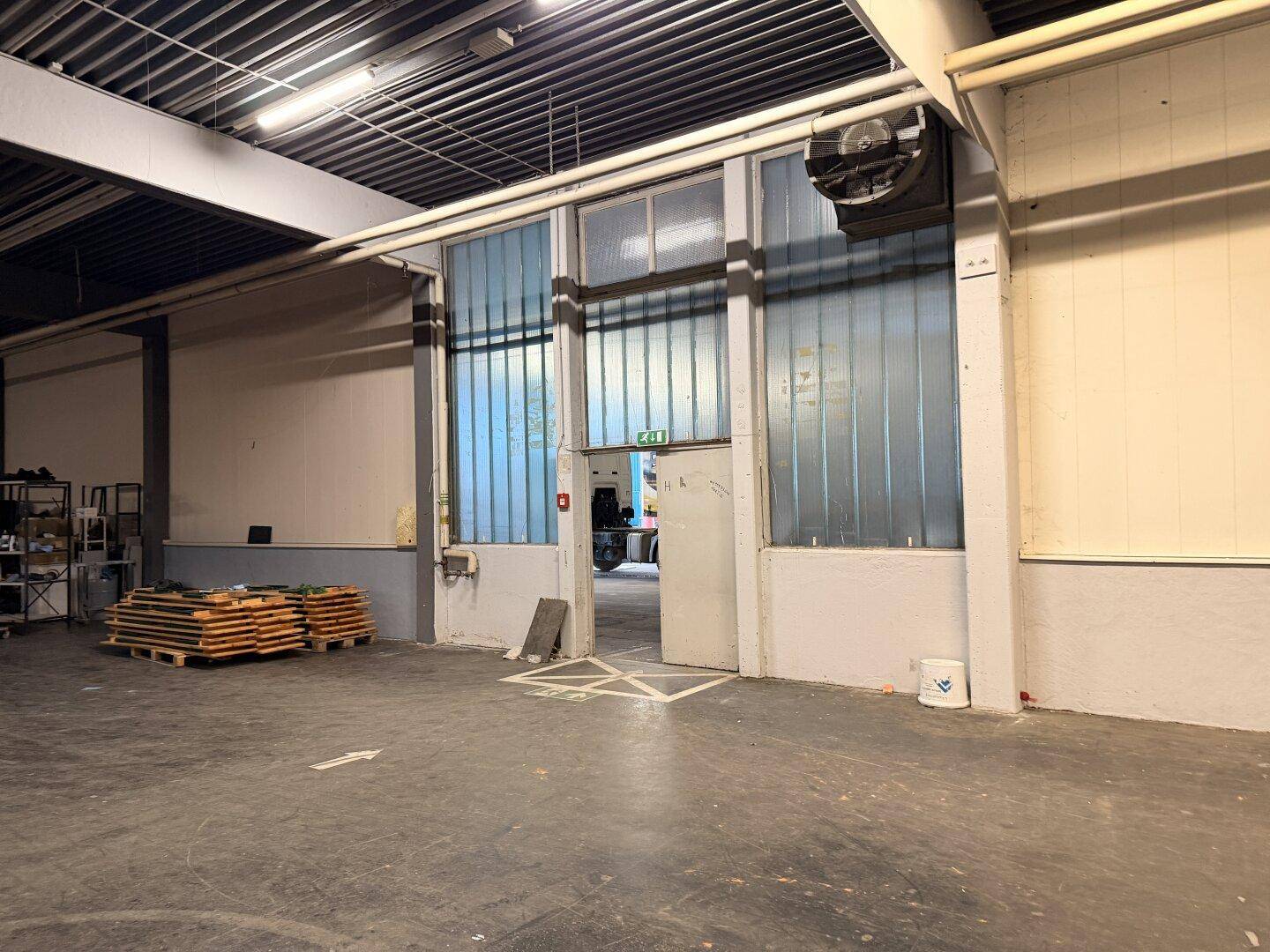 Eingangsbereich eines Lagerraums mit großen Türen, Glasbausteinen und einem Wandventilator für Belüftung.