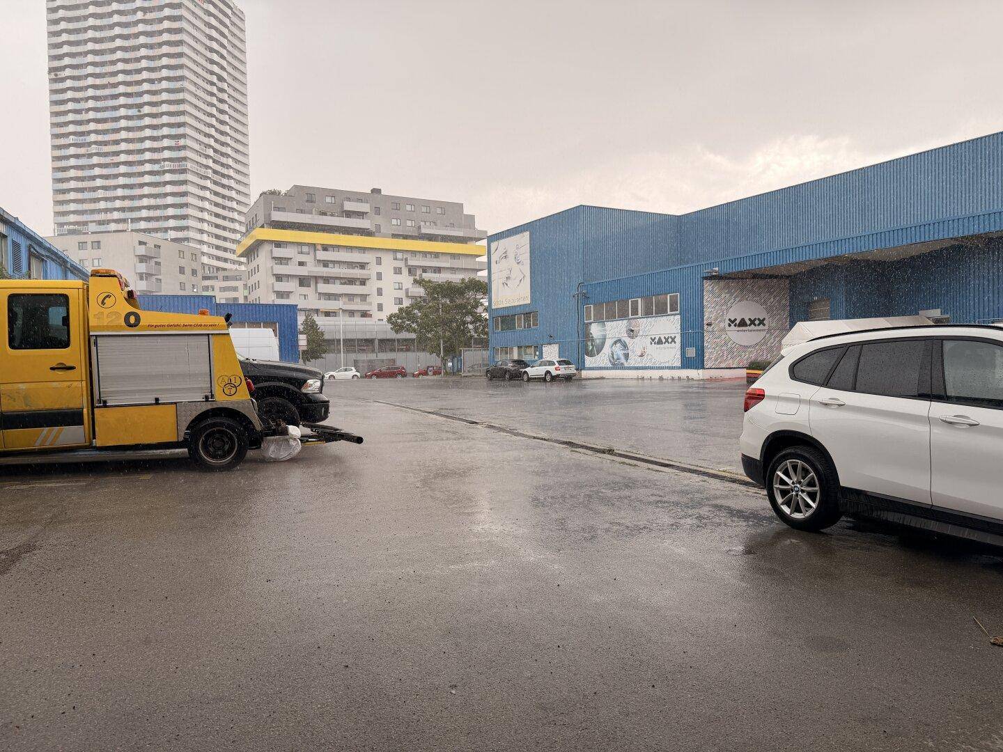 Außenansicht eines Industriegebiets mit blauen Gebäuden, einem Hochhaus und geparkten Autos bei Regenwetter.