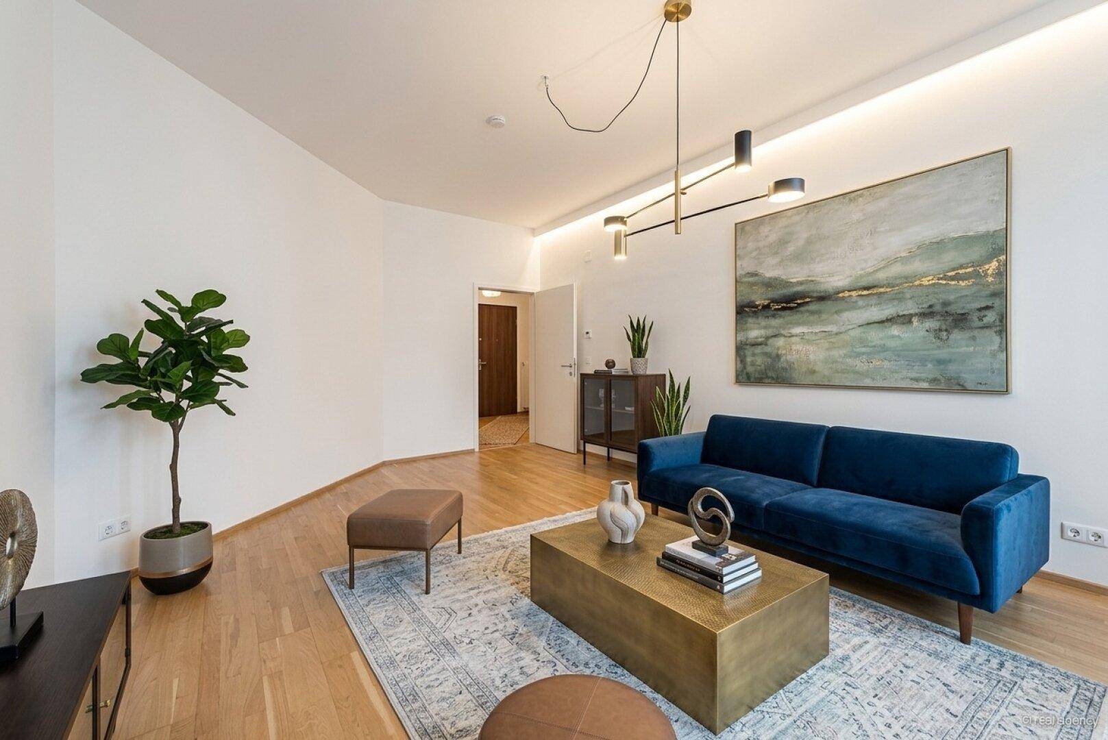 Modernes Wohnzimmer mit blauem Samtsofa, hellem Holzboden und stilvoller Deckenbeleuchtung, ergänzt durch ein abstraktes Kunstwerk.