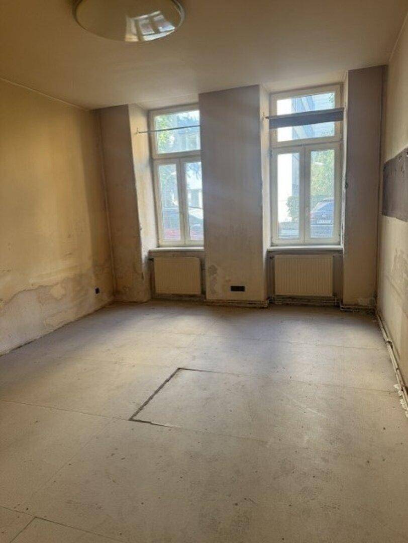 Unmöblierter Raum in einem Altbau mit zwei Fenstern und sichtbarem Betonboden.