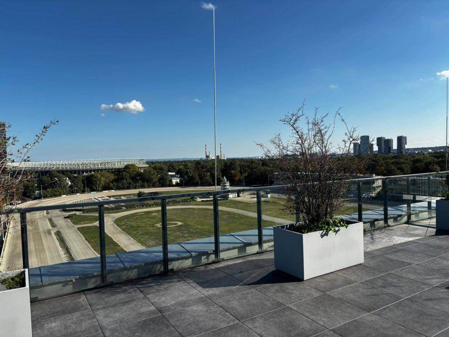 Weitläufige Dachterrasse mit Bepflanzung und Blick auf ein Stadion sowie die Stadtsilhouette.