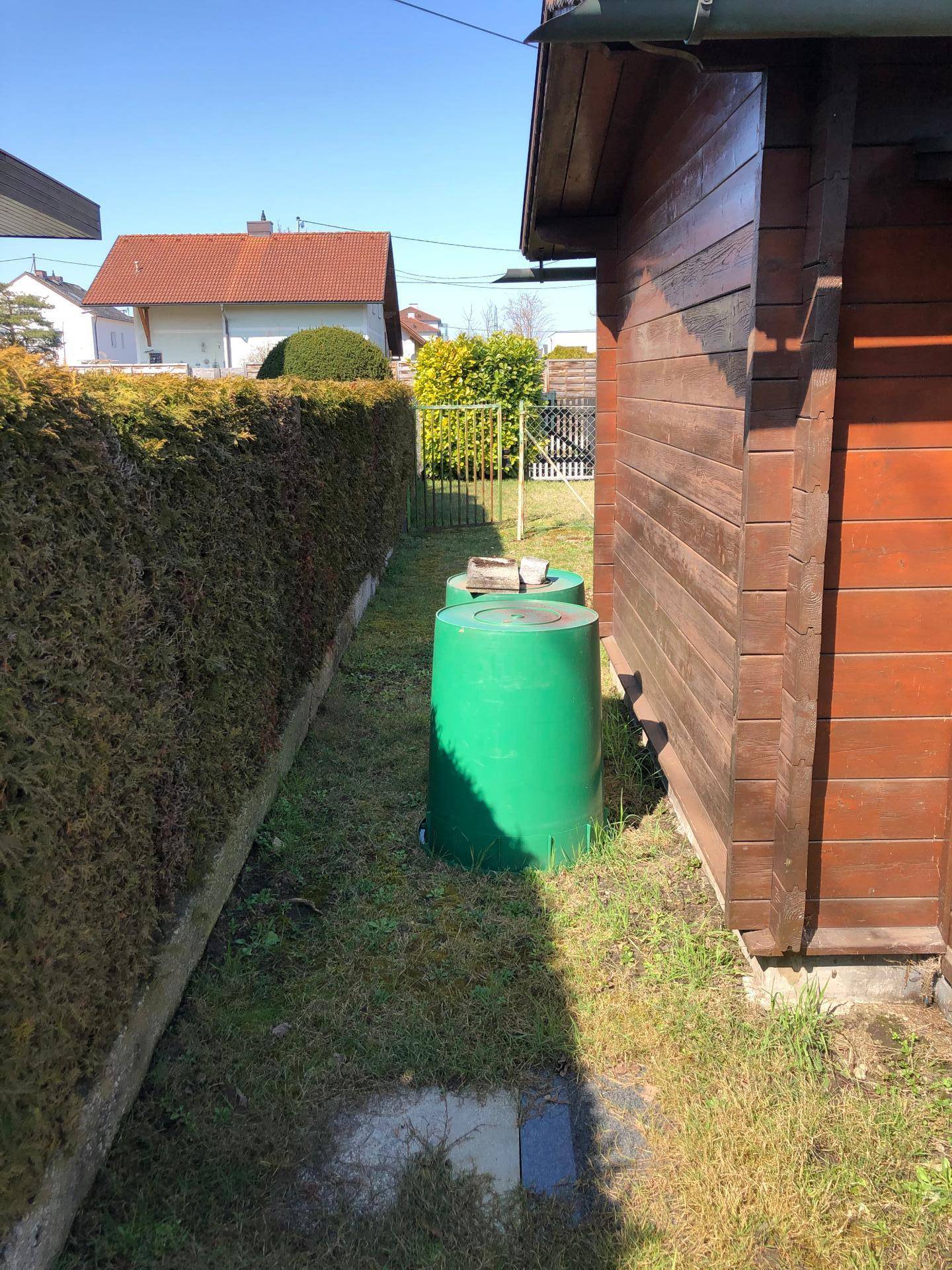 Seitenansicht des Holzschuppens mit grünen Regentonnen und einer hohen Hecke.