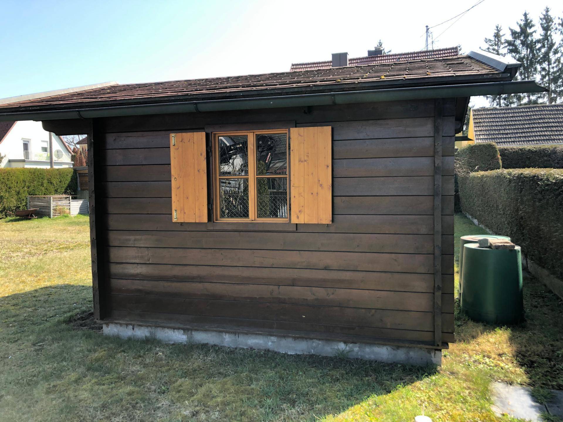Holzschuppen mit kleinen Fenstern und Fensterläden, umgeben von Rasen und Hecke.