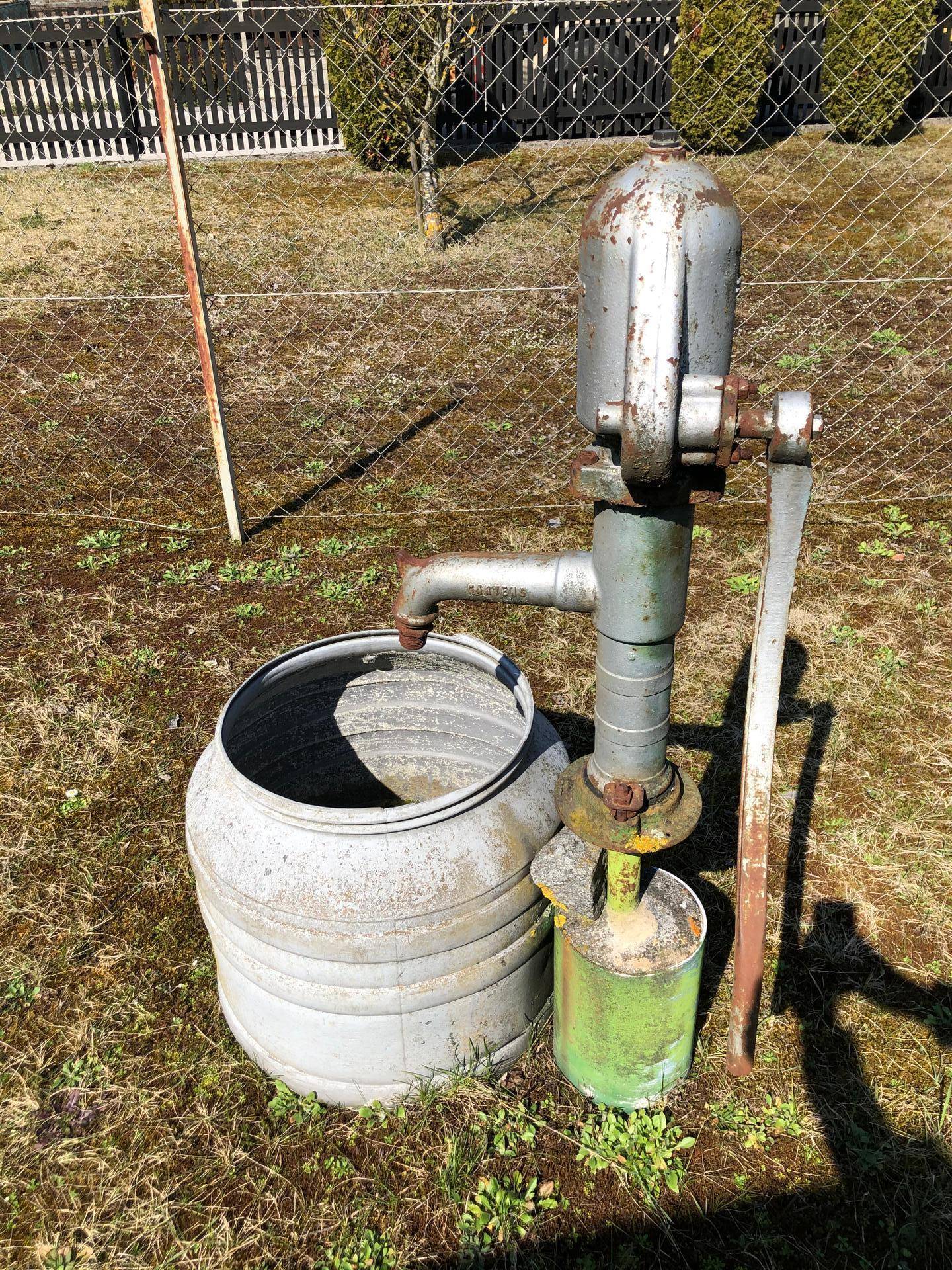 Alte Wasserpumpe und Metallfass im Garten, umgeben von Gras und Zaun.