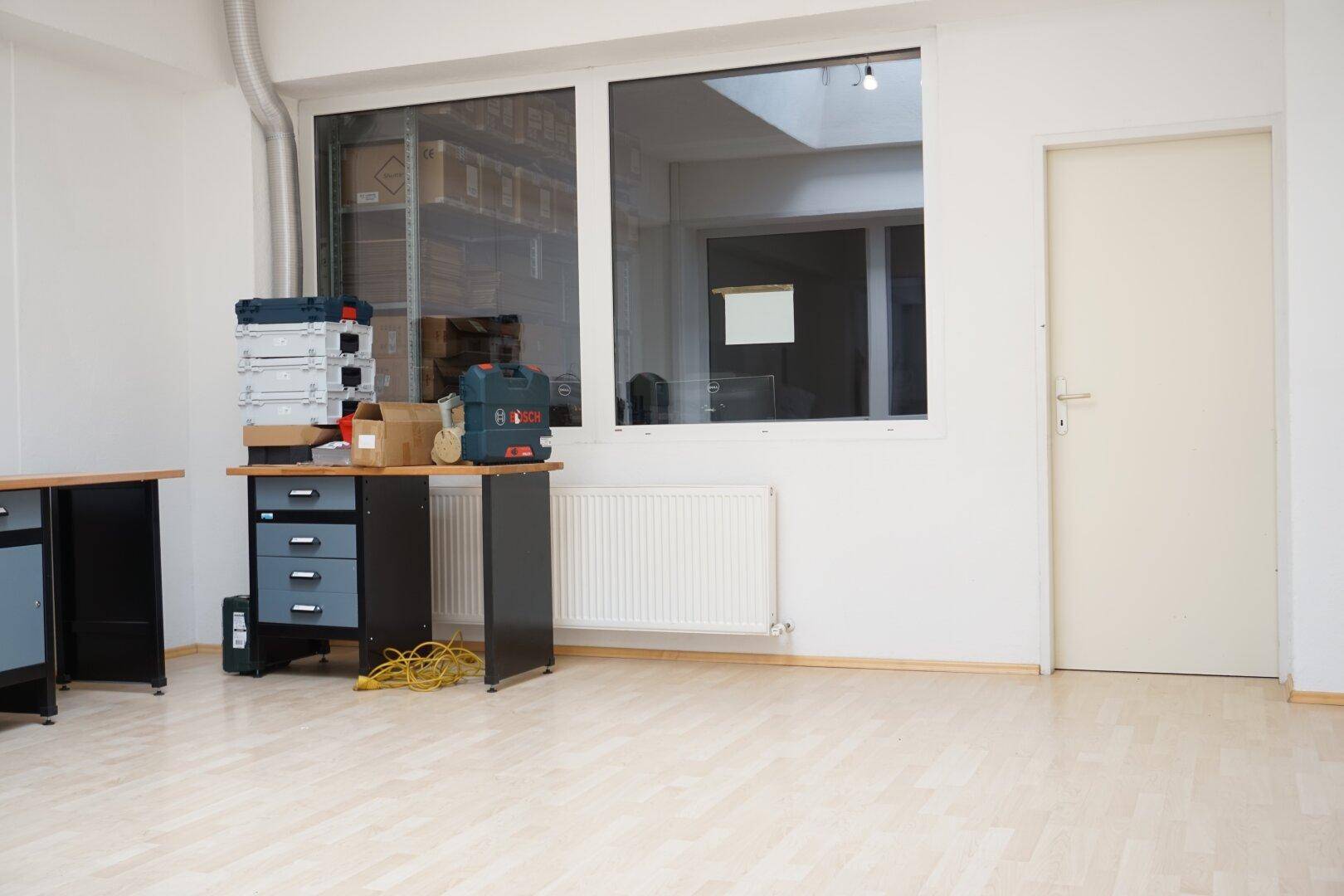 Heller Werkstattraum mit Holzboden, großem Fenster und Arbeitsbereich, ideal für handwerkliche Tätigkeiten oder Lagerung.